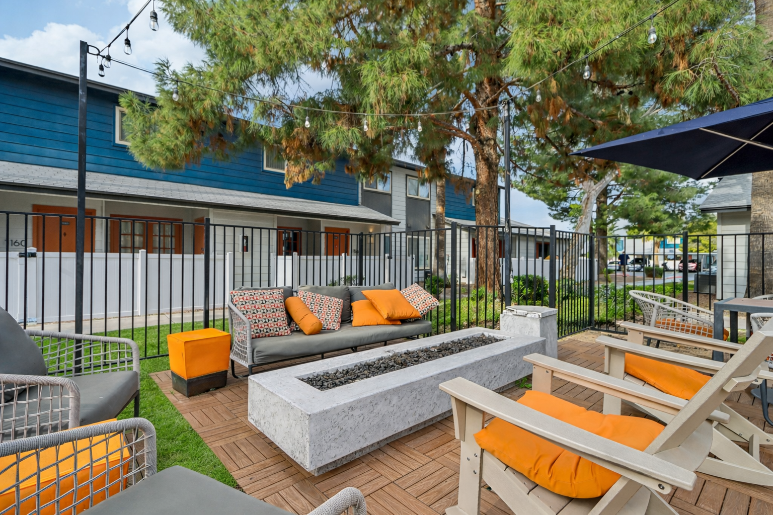 Outdoor lounge area featuring comfortable seating with orange cushions, a modern stone fire pit, and greenery. A fence surrounds the space, with string lights overhead, creating a cozy atmosphere. The background includes a building with blue and gray exteriors.