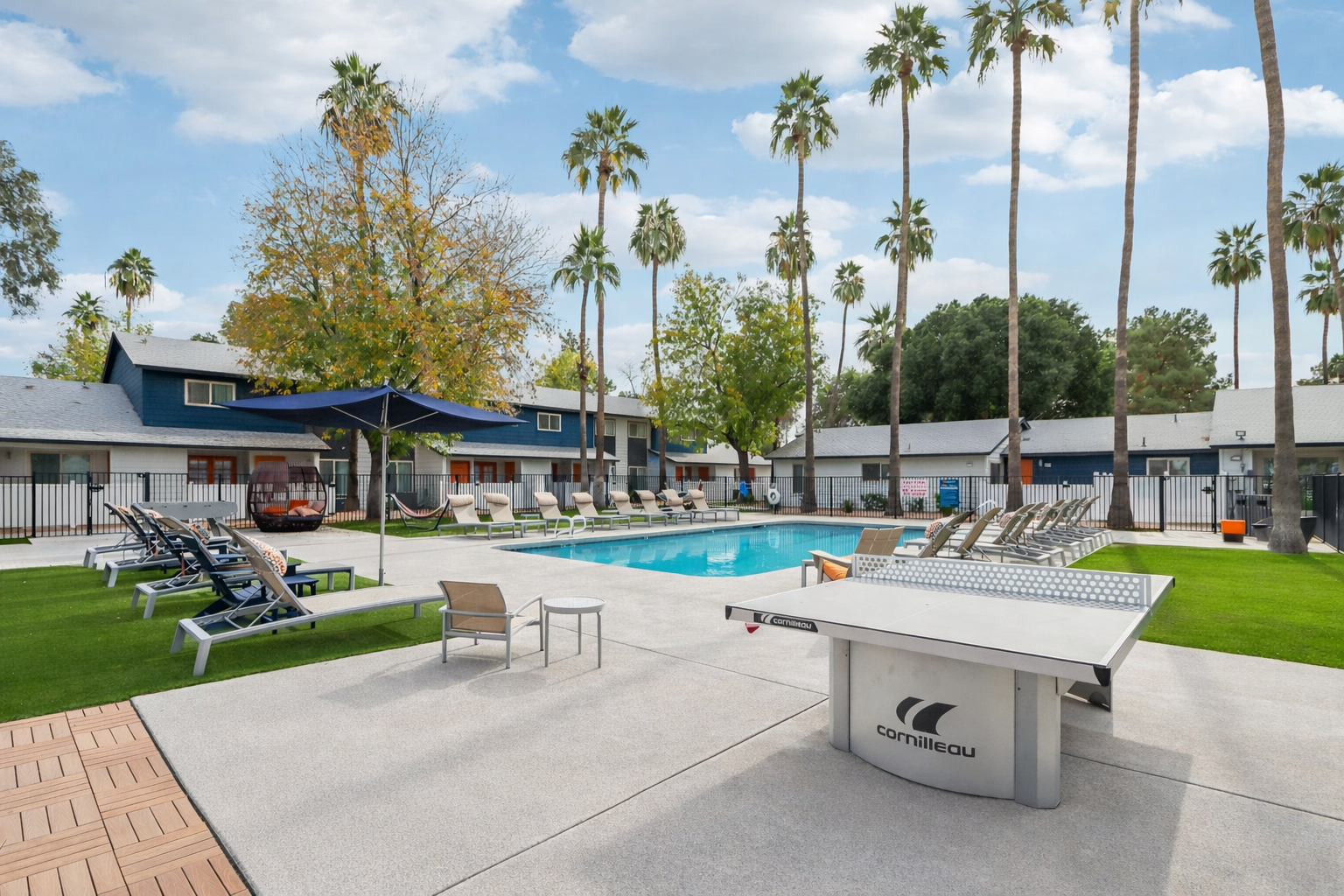 A courtyard featuring a swimming pool surrounded by lounge chairs and palm trees. In the foreground, there's a table tennis setup. The background shows multi-story buildings with a blue sky and scattered clouds above. The area is well-maintained with green grass and a relaxing atmosphere.