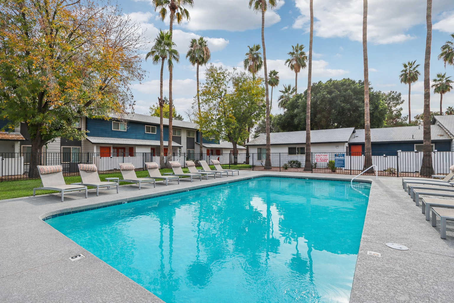 A clear blue swimming pool surrounded by lounge chairs, with tall palm trees and colorful autumn foliage in the background. The pool reflects the sky and nearby buildings, creating a tranquil outdoor setting perfect for relaxation.