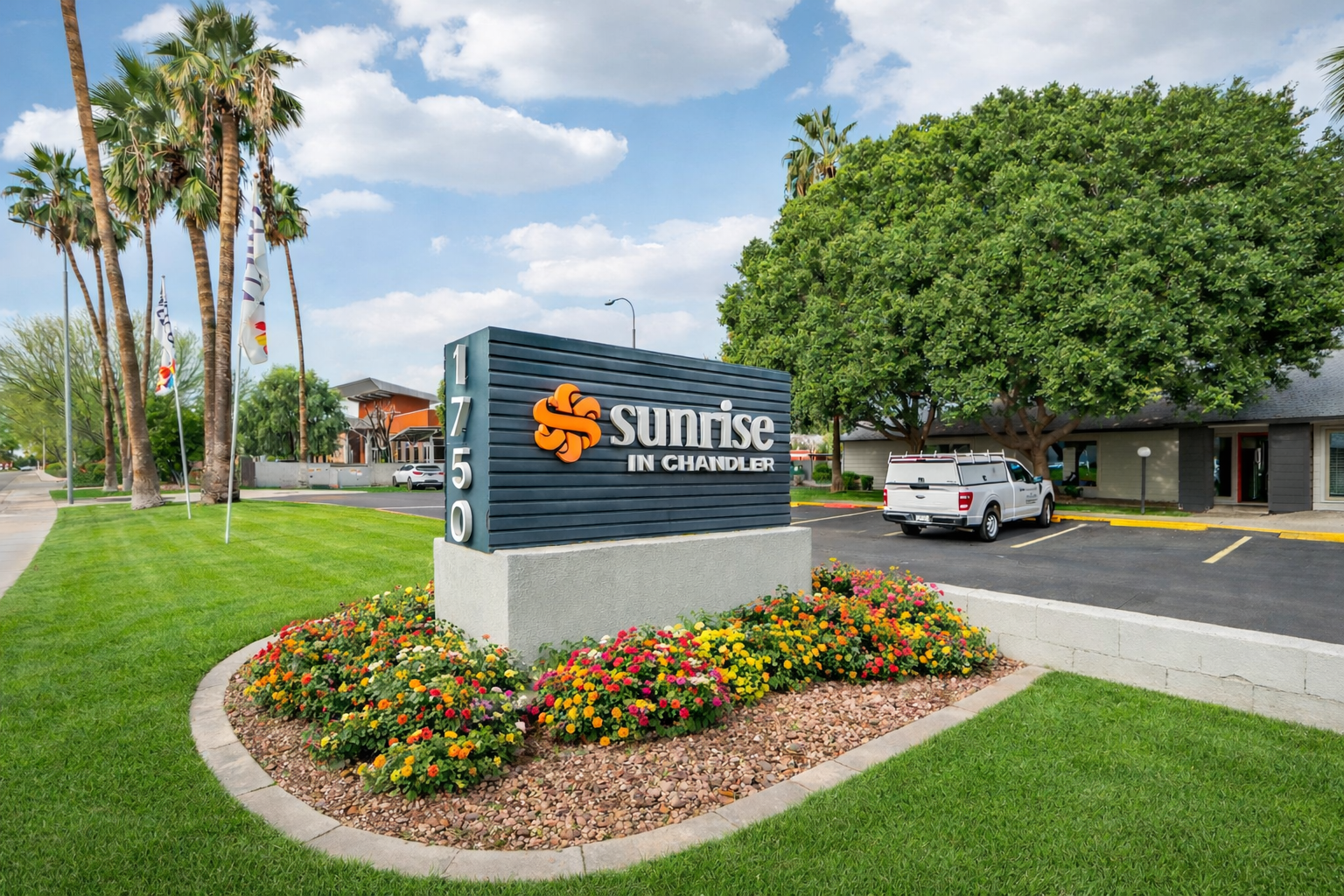 Sign for "Sunrise in Chandler" prominently displayed in front of a complex. The sign features an orange logo and is surrounded by well-maintained landscaping with colorful flowers and palm trees. The area has a paved parking lot and a building in the background under a partly cloudy sky.