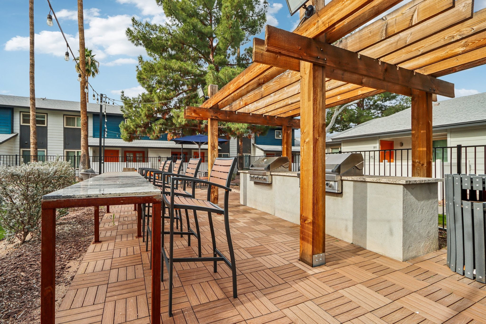 Outdoor grilling area featuring a wooden pergola with bar-style seating, stone countertops, and built-in grills. Surrounding the space are landscaped areas and a mix of multi-colored buildings in the background. The sky is clear with a few clouds, creating a bright and inviting atmosphere.