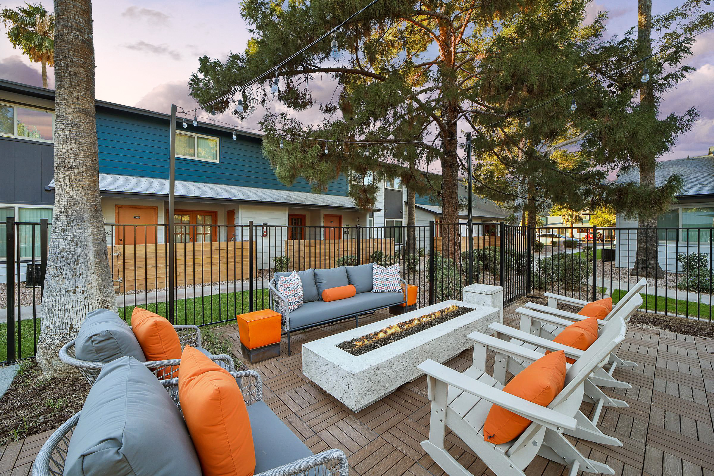 A cozy outdoor lounge area featuring a concrete fire pit surrounded by white adirondack chairs and a gray sofa with orange pillows. The setting is framed by tall trees and includes modern buildings in the background, creating a welcoming atmosphere for relaxation.
