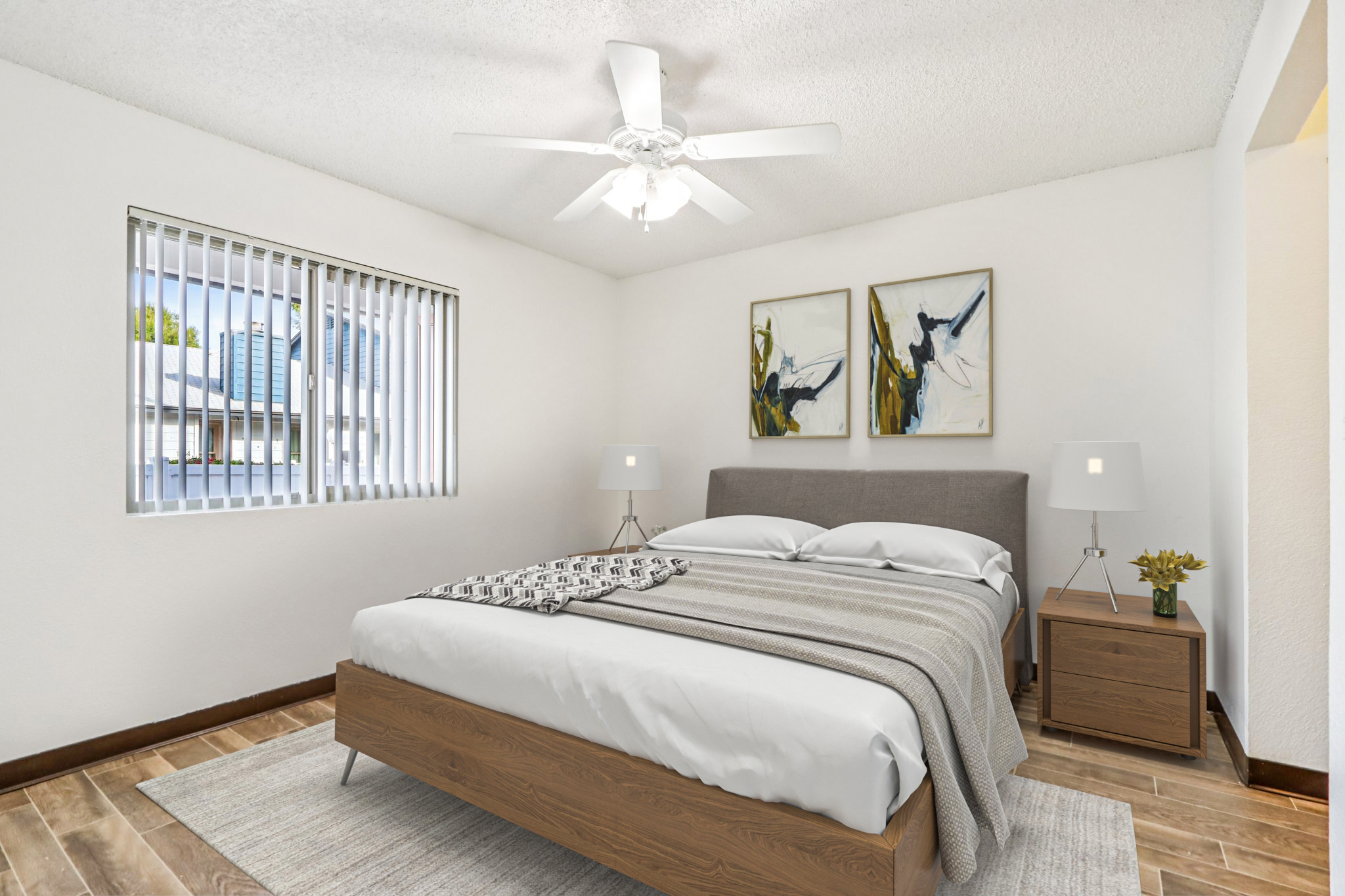 A neatly arranged bedroom featuring a queen-sized bed with a gray blanket, two modern lamps on nightstands, and abstract paintings on the wall. A ceiling fan is above, and a window with vertical blinds allows natural light to enter. The floor is covered with light wood planks and a neutral rug.