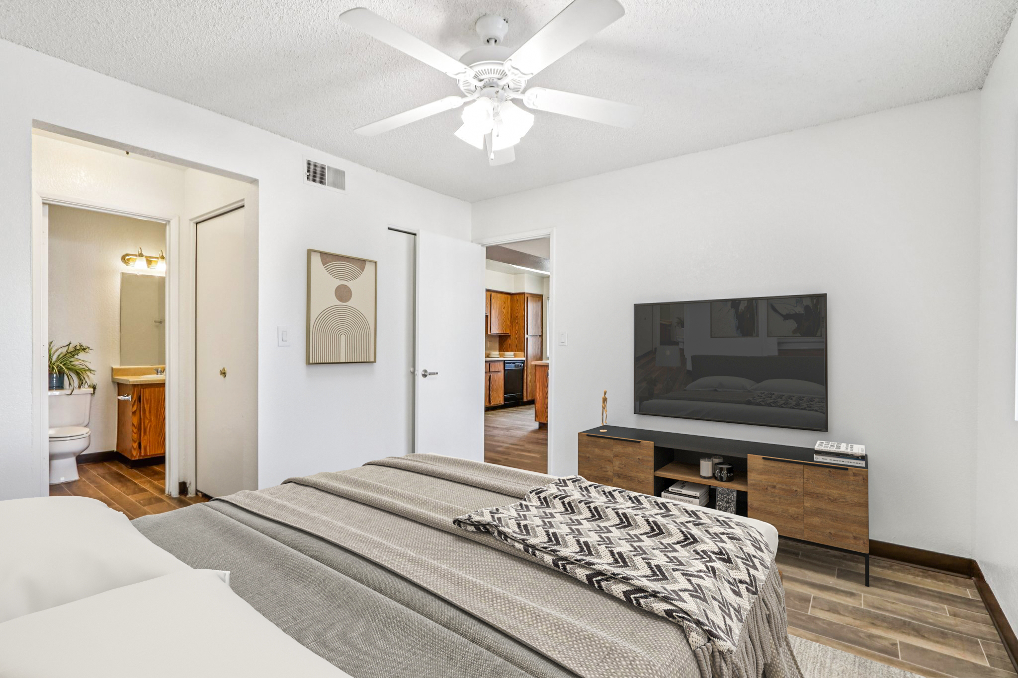 A cozy bedroom featuring a neatly made bed with a patterned throw blanket. There’s a ceiling fan, a large TV on a wooden dresser, and light-colored walls. A doorway leads to a bathroom, and another door opens to a kitchen area. The floor has a wooden finish, enhancing the warm atmosphere of the space.
