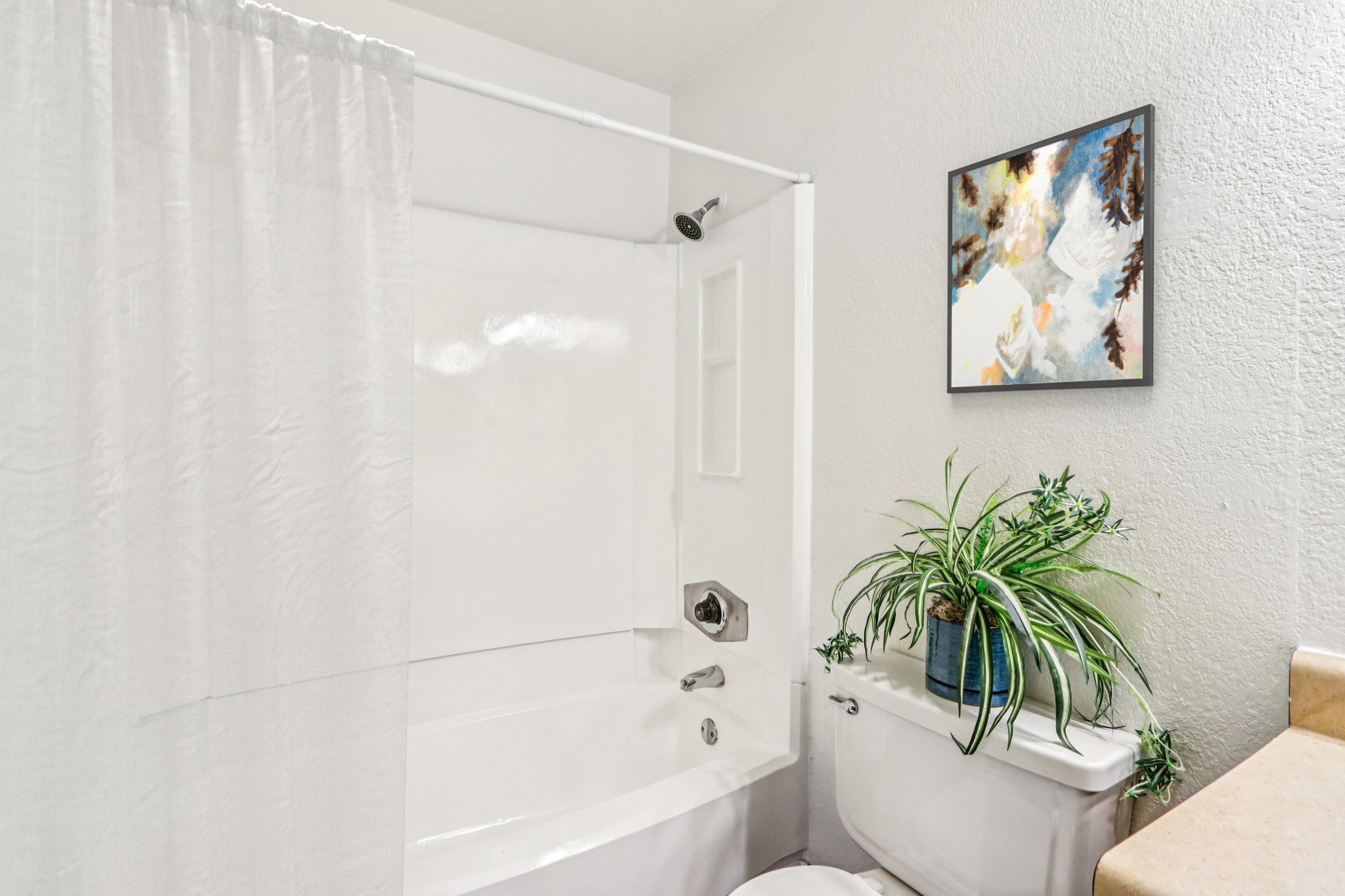 A bright bathroom featuring a white tub with a shower curtain, a modern piece of wall art, a green potted plant on a counter, and a toilet. The walls are light-colored, creating an airy feel in the space.