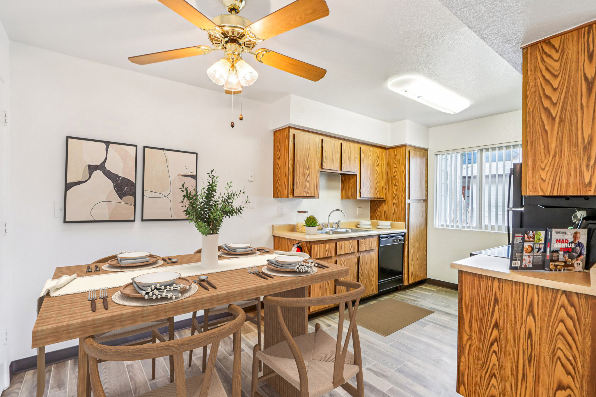 A modern kitchen and dining area featuring wooden cabinets, a round dining table set for four, and a ceiling fan. The space includes a black dishwasher, light-colored walls, and large windows providing natural light. Decorative elements like wall art and a potted plant enhance the aesthetic.