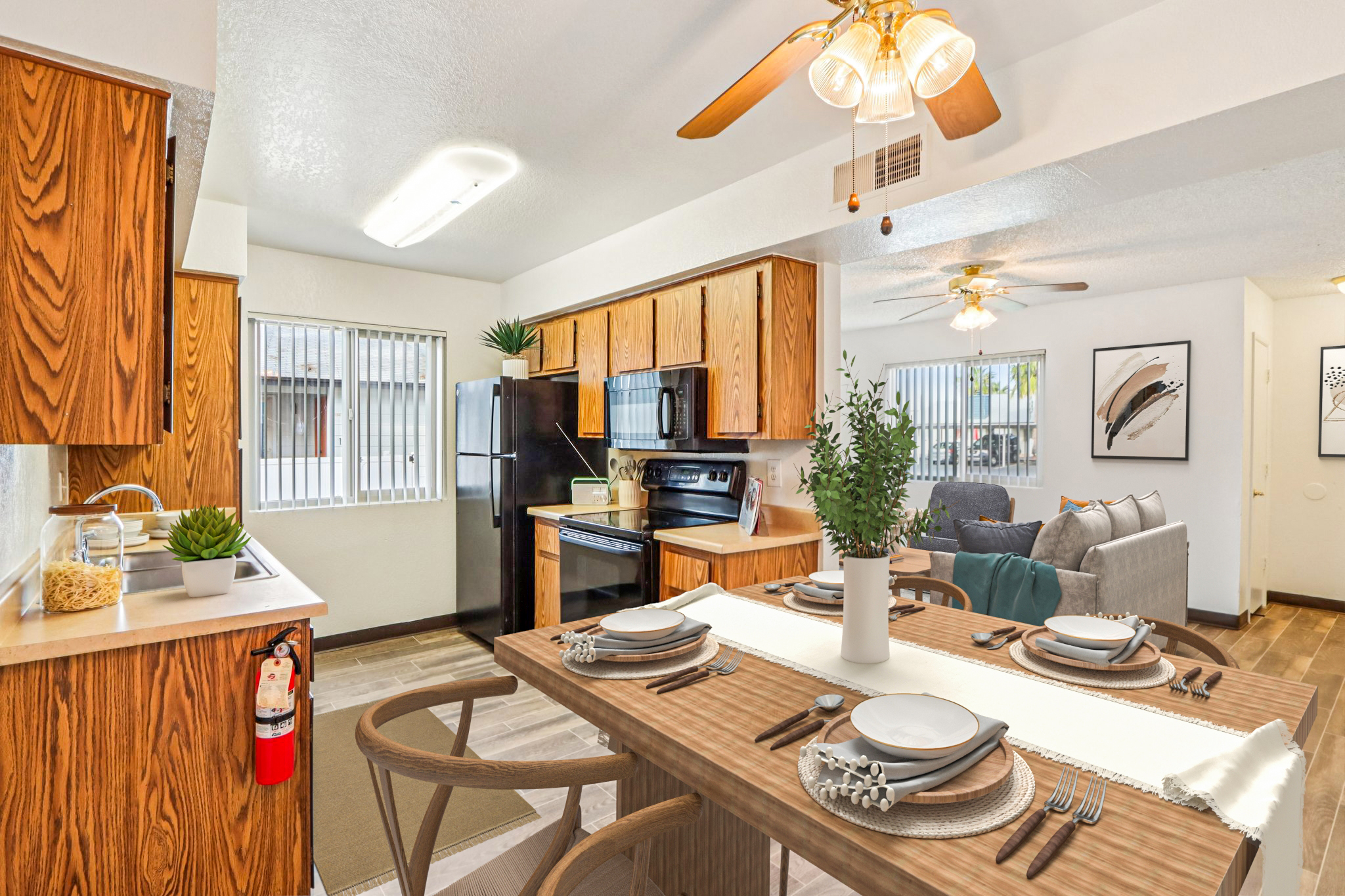 A modern kitchen and dining area featuring wooden cabinetry, a black refrigerator, and stainless-steel appliances. A dining table is set with dishware and a centerpiece, with a cozy living area visible in the background. Large windows bring in natural light, enhancing the bright and inviting atmosphere.