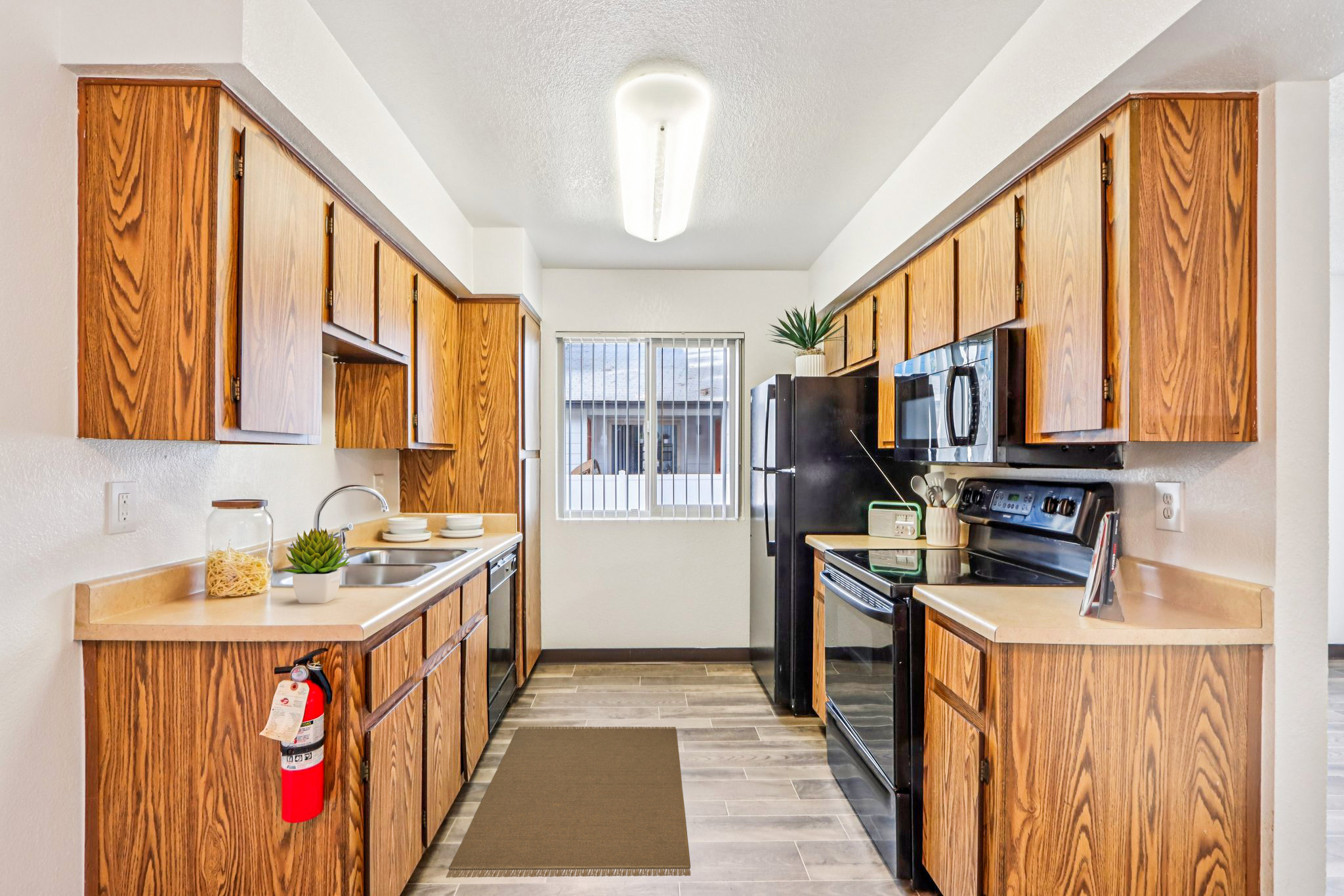 A modern kitchen featuring wooden cabinets, a stainless steel sink, and an electric stove. There is a black refrigerator, a window with vertical blinds, and a small rug on the floor. Countertops hold plates and a jar of pasta, while a potted plant adds a touch of greenery.