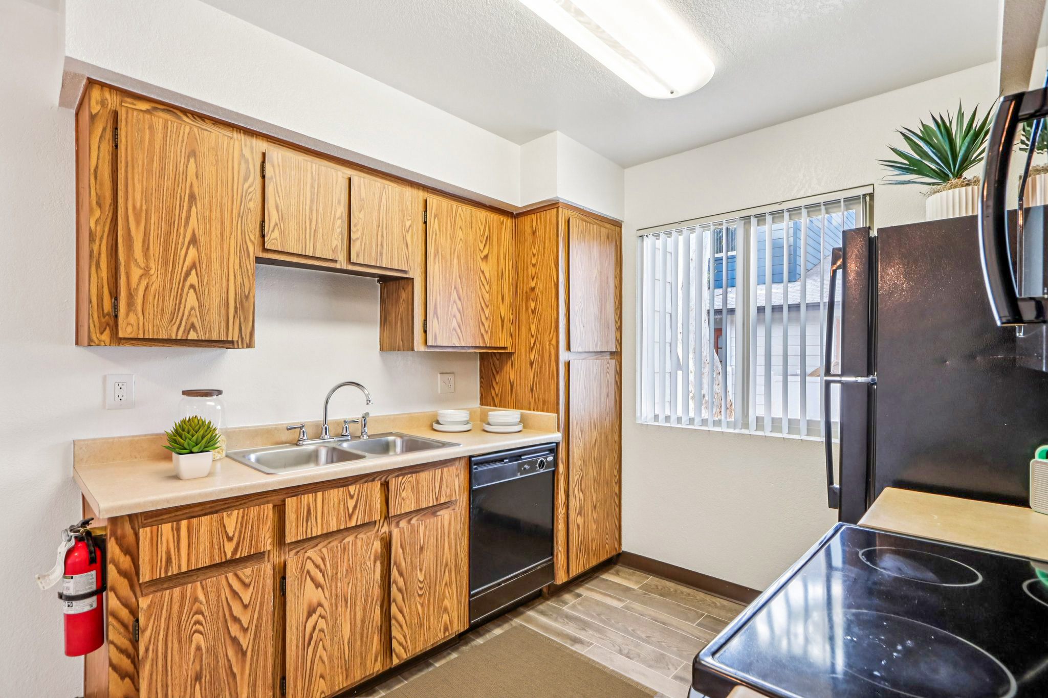A modern kitchen featuring wooden cabinets, a double sink, and black appliances, including a dishwasher and stove. Natural light enters through a window with blinds. A small plant decorates the countertop. A fire extinguisher is mounted on the wall for safety. The overall design is functional and inviting.