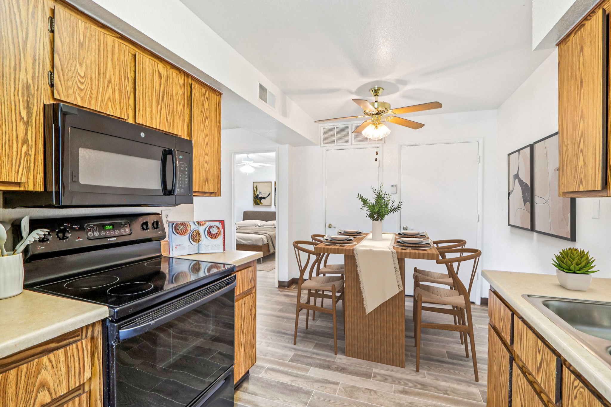 A bright, modern kitchen with wooden cabinetry and stainless steel appliances. A dining area features a wooden table set for four with a centerpiece. The space has light-colored walls, a ceiling fan, and natural light coming from nearby windows, creating an inviting atmosphere.