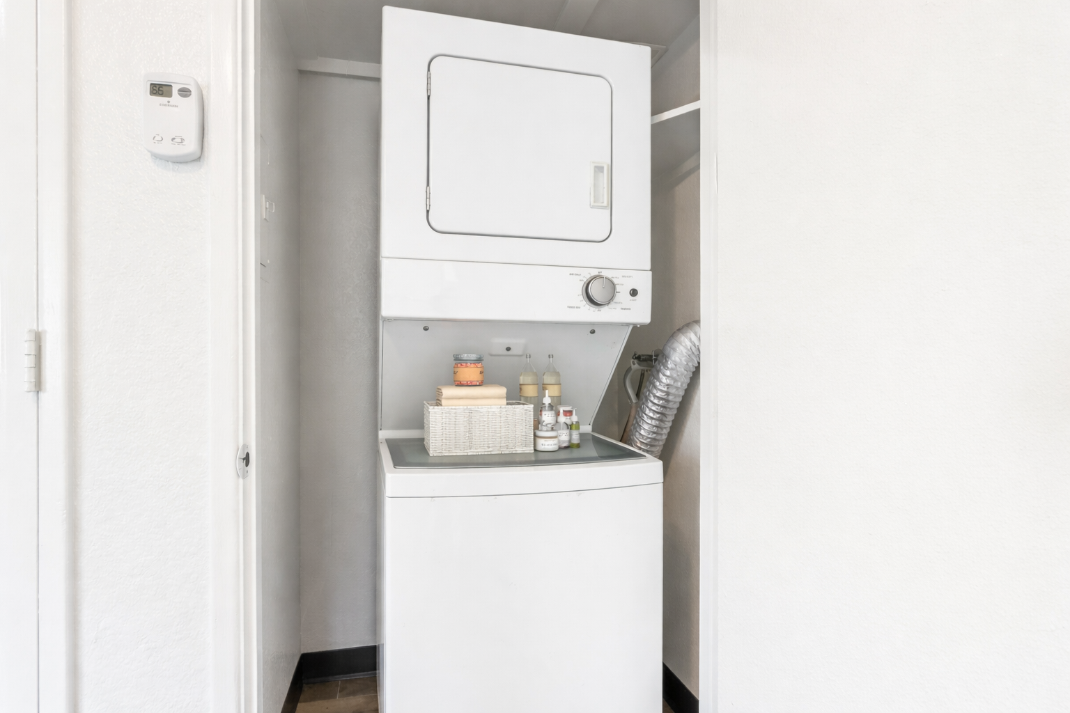 A compact laundry area featuring a stacked washer and dryer in a small closet space. On top of the washer, there is a white storage basket and neatly arranged bottles. The walls are painted white, contributing to a clean, organized appearance. An air duct is visible, indicating ventilation.