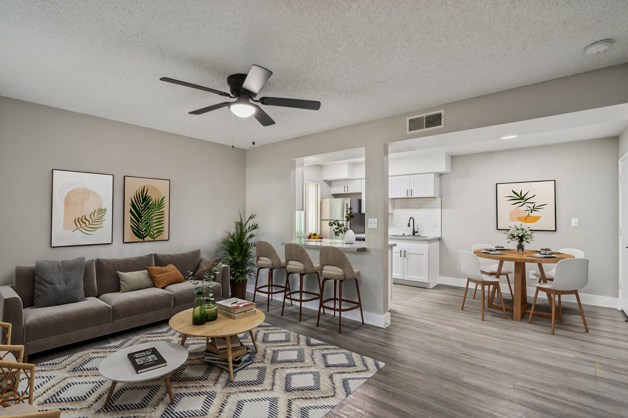 A modern living space featuring a cozy sofa, stylish wall art, and a round dining table with chairs. The open layout connects the living area to a kitchen with white cabinets. Potted plants add a touch of greenery, while a light wood floor enhances the contemporary feel of the room.