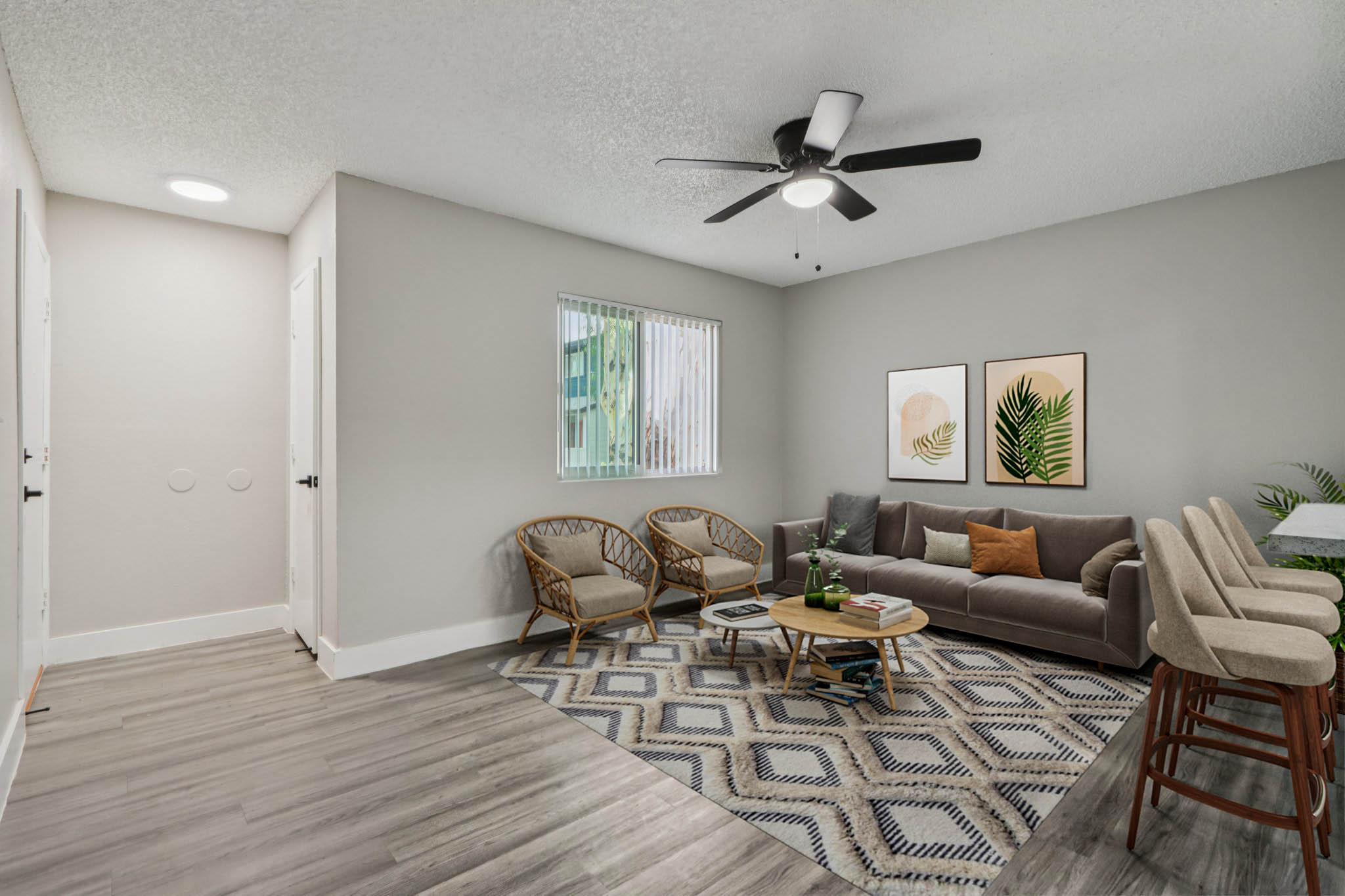 A modern living room featuring a gray sofa, two wicker armchairs, a round coffee table with decorative plants, and a geometric area rug. The walls are light gray, with large windows allowing natural light. Two framed botanical prints hang on the wall, and there is a dining area with bar stools in the background.