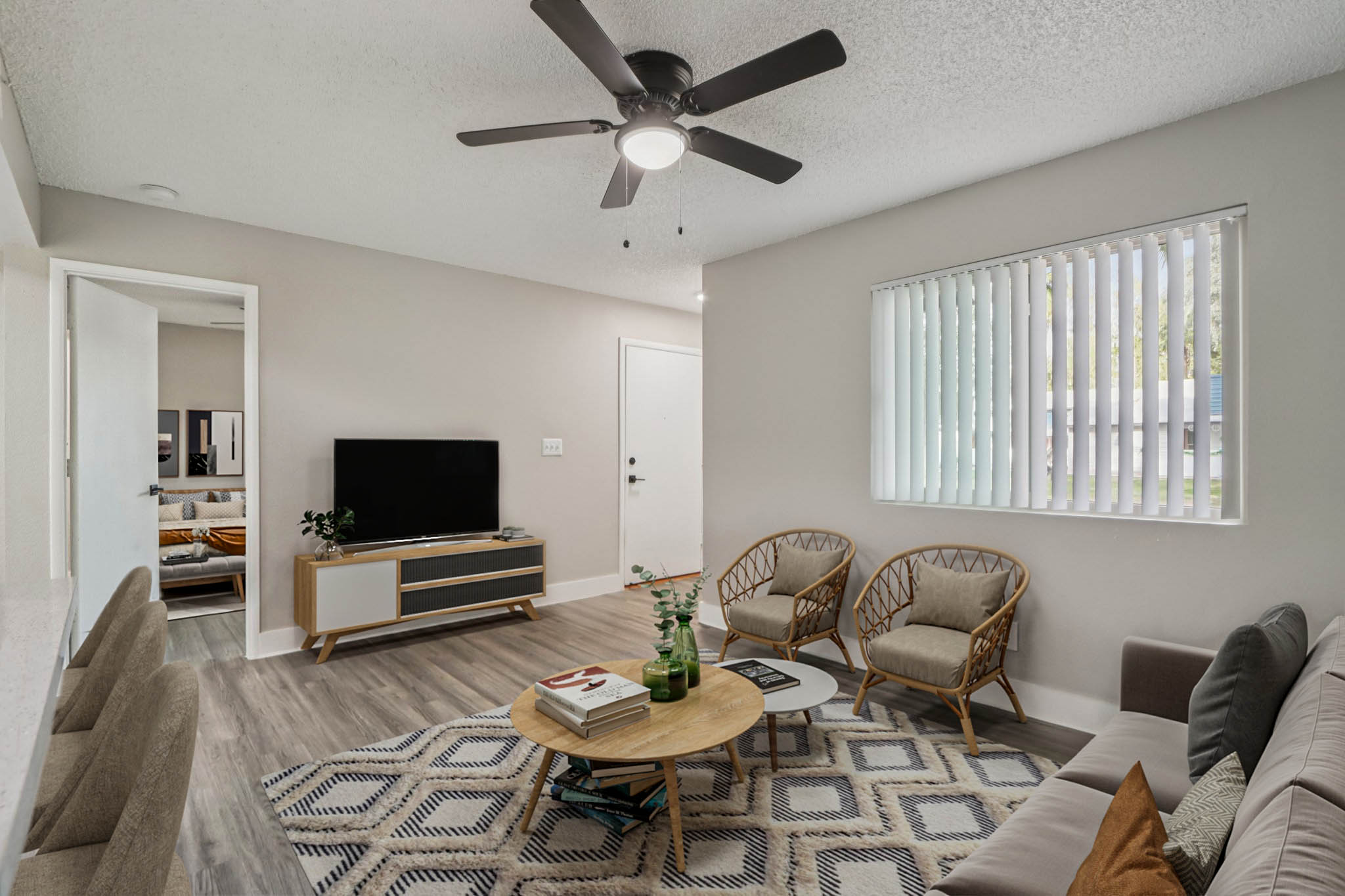 A modern living room featuring a light color scheme, a ceiling fan, a mounted TV on a wooden console, and a round coffee table with books and a plant. There are two styled chairs and a sofa, with large windows allowing natural light. A doorway in the background leads to another room.