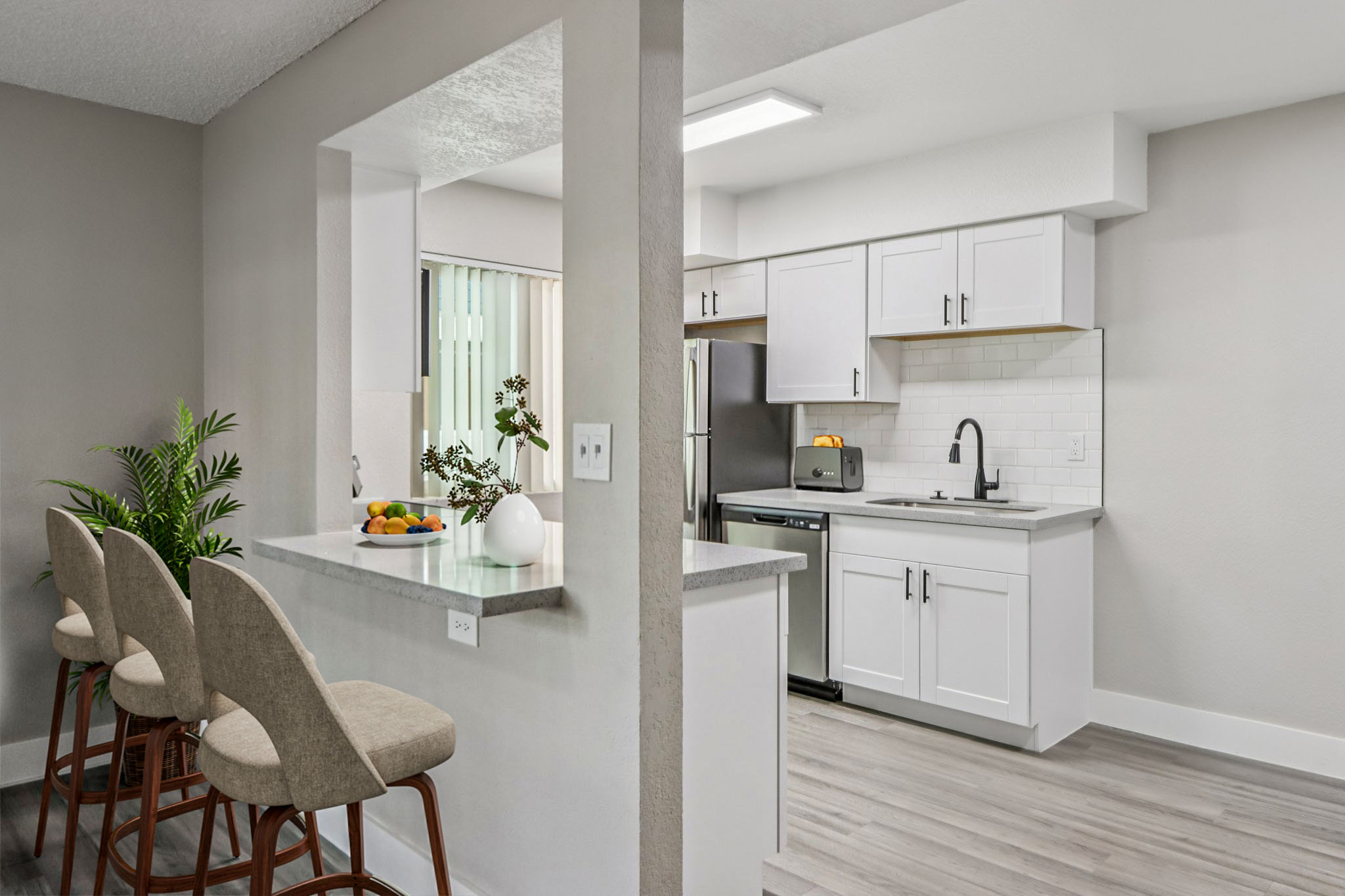 Modern kitchen featuring white cabinetry, a patterned backsplash, and stainless steel appliances. A small island with three barstools separates the kitchen from a living area, which includes a bowl of fruit and a plant. Natural light streams in through a window, highlighting the clean, minimalistic design.