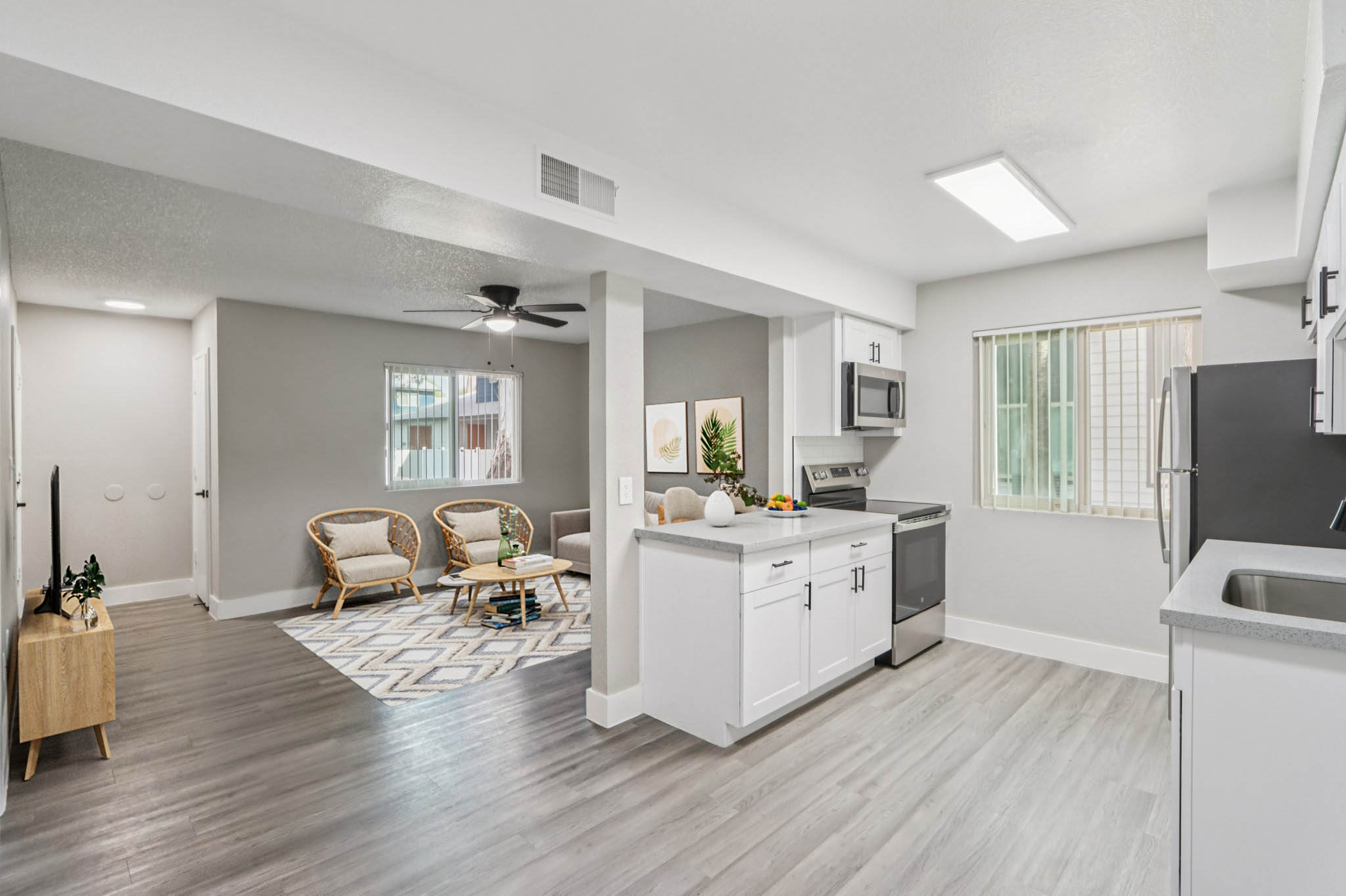 Modern open-concept kitchen and living area featuring a sleek kitchen with stainless steel appliances, white cabinetry, and a dining space. The living room includes two rattan chairs and a patterned rug, with natural light streaming through a window. Neutral-toned walls and flooring create a spacious, inviting atmosphere.