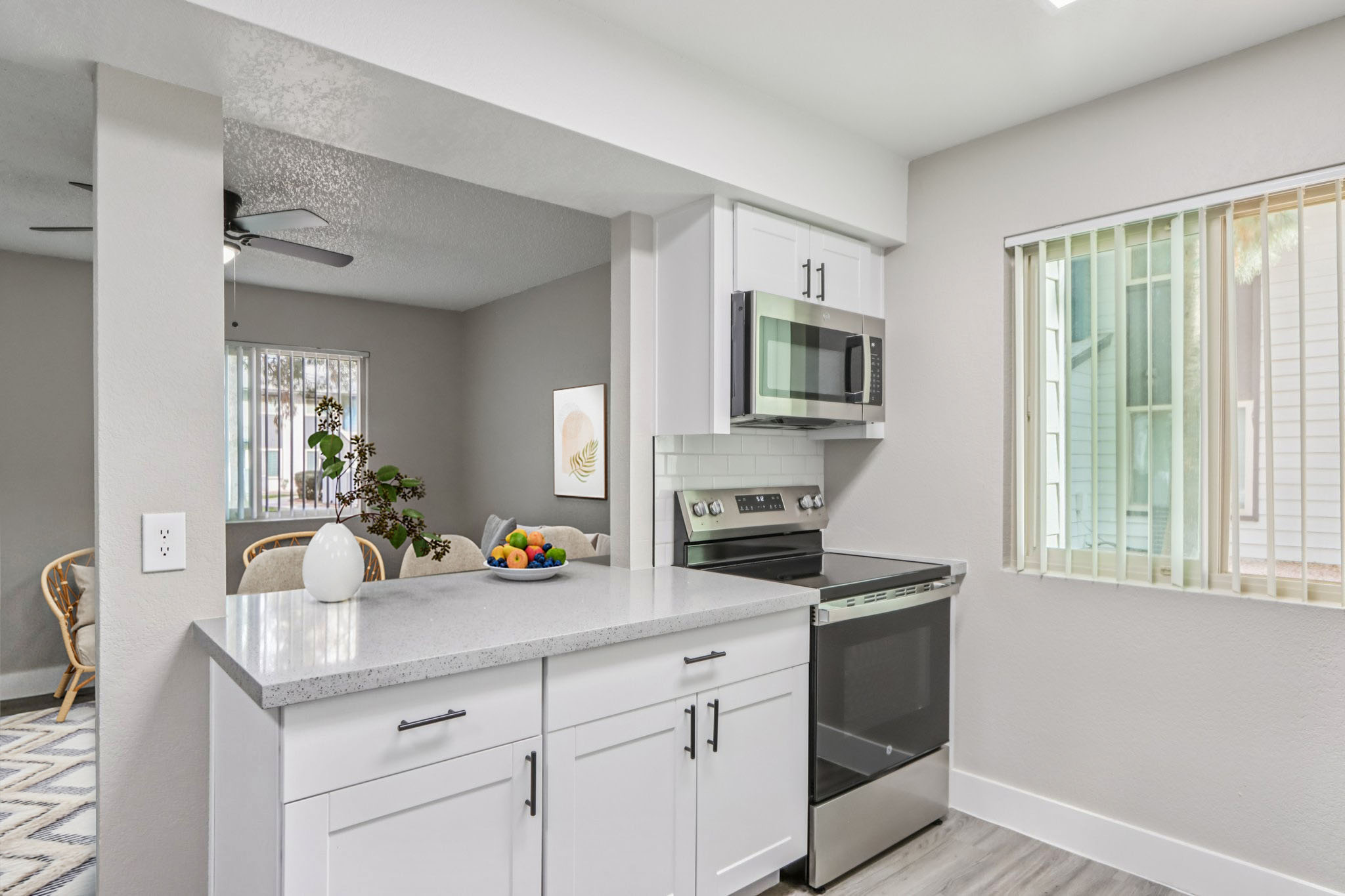 Modern kitchen featuring white cabinetry, a stainless steel oven and microwave, a light-colored countertop, and a bowl of fresh fruit. The space is well-lit with natural light filtering through a nearby window, and a decorative plant can be seen in the background.