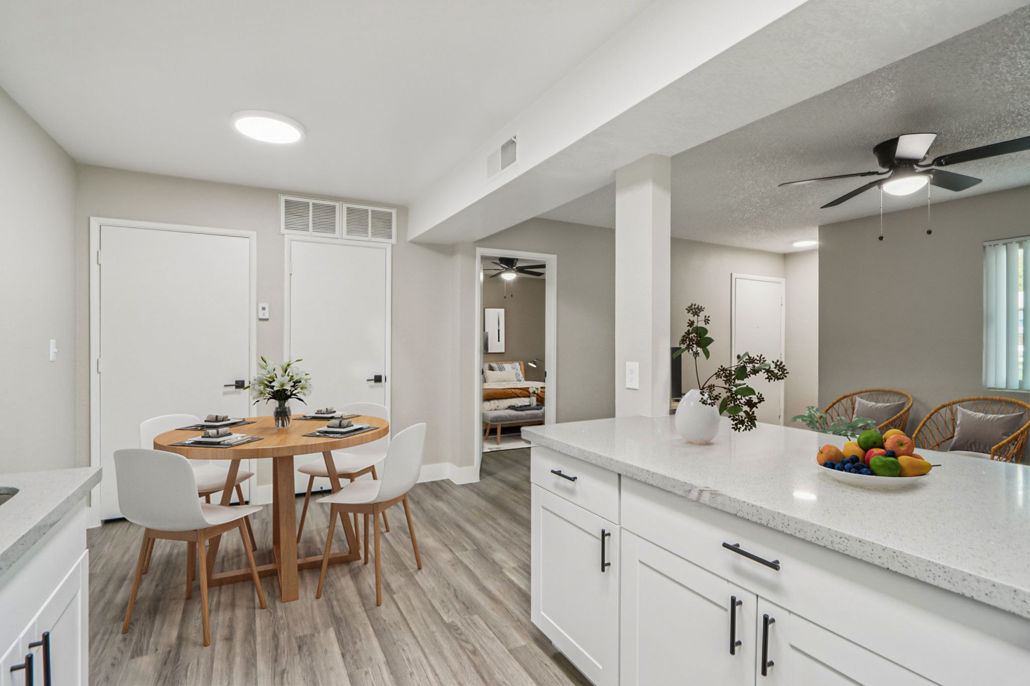 A bright and modern kitchen and dining area featuring a round wooden table with four white chairs, a vase of flowers, and a bowl of fruits. The open layout includes a view into a cozy living space with rattan chairs and a fan, showcasing light gray walls and wooden flooring.