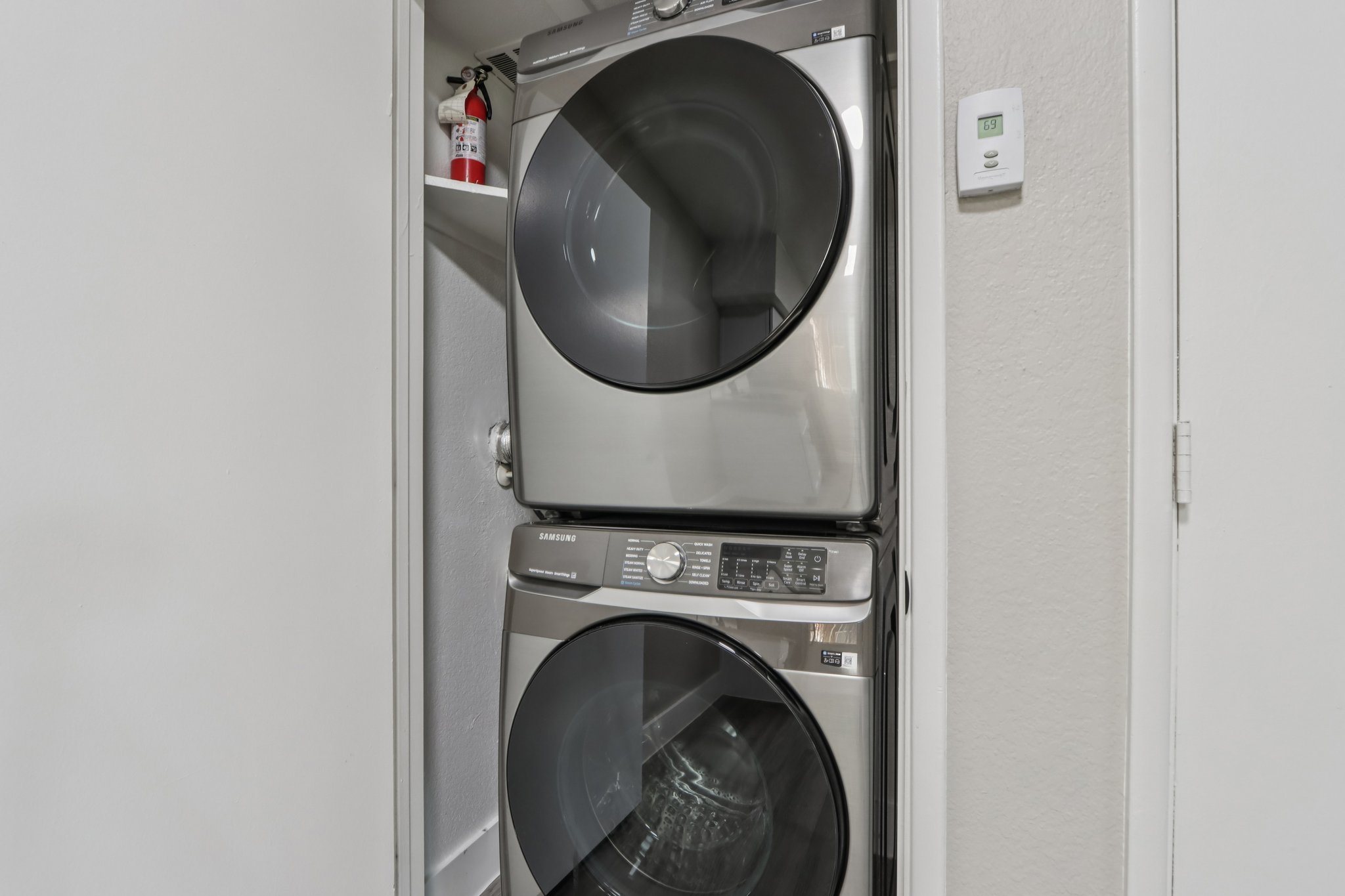 A stackable washer and dryer set in a narrow laundry space, featuring a silver finish. A fire extinguisher is visible on a shelf nearby, alongside a wall-mounted device displaying temperature controls. The walls are painted in a light color, enhancing the brightness of the area.