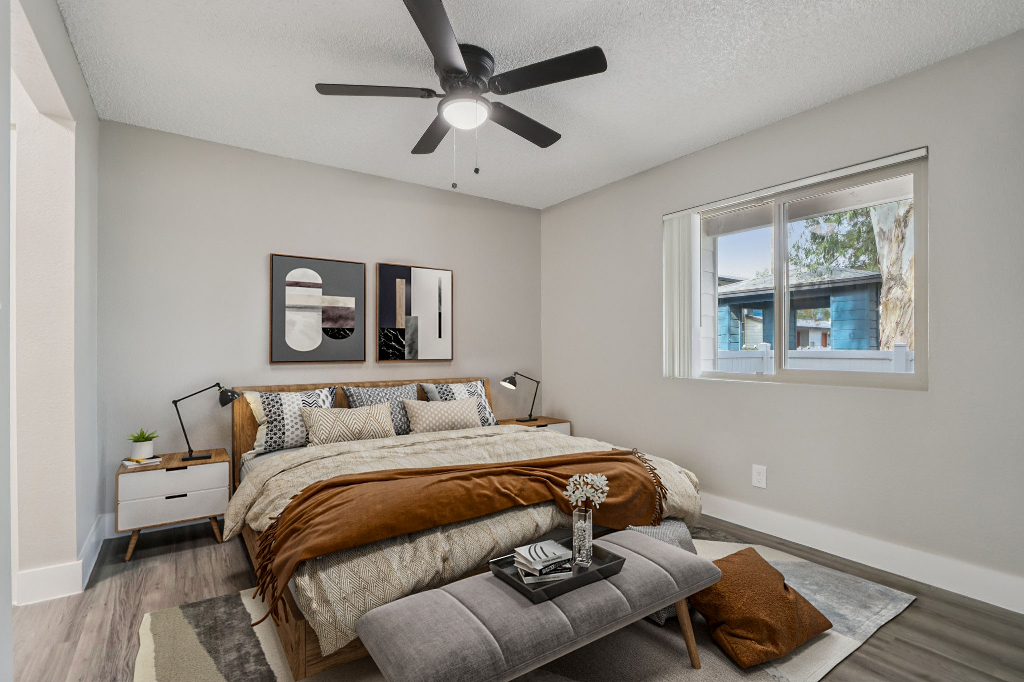 A modern bedroom featuring a neatly made bed with decorative pillows and a cozy throw. The room includes a fan, a nightstand with decorative items, and large windows that bring in natural light. Neutral-colored walls and a stylish rug enhance the contemporary aesthetic.