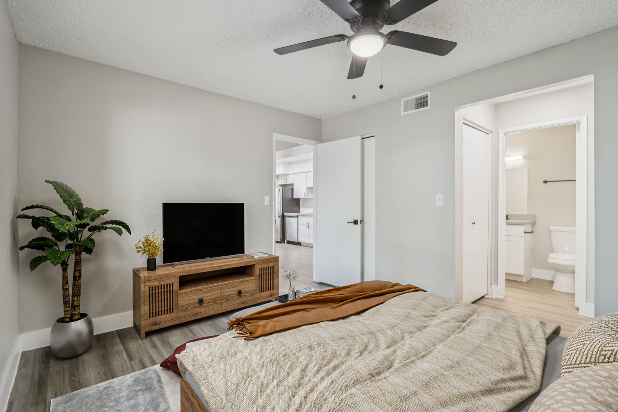 A cozy bedroom featuring a large bed with a textured blanket, a wooden TV stand with a television, and a potted plant. In the background, a bathroom door is visible alongside a door leading to another room, with neutral-colored walls and a ceiling fan. Natural light brightens the space.