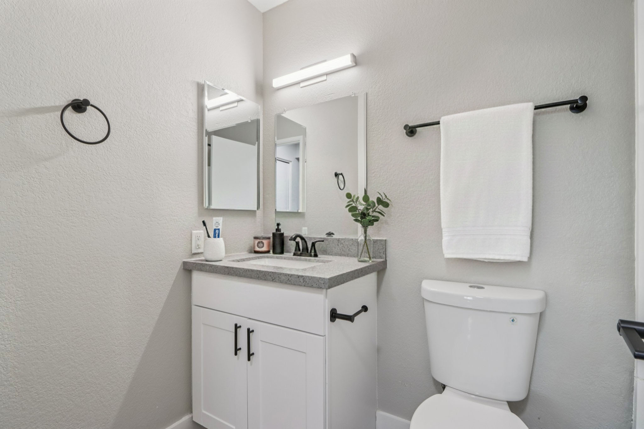 A modern bathroom with a neutral color scheme. It features a compact vanity with gray countertop, two mirrors, a black towel rack holding a white towel, and a small potted plant. A white toilet is positioned beside the vanity, adding to the clean and minimalist aesthetic.