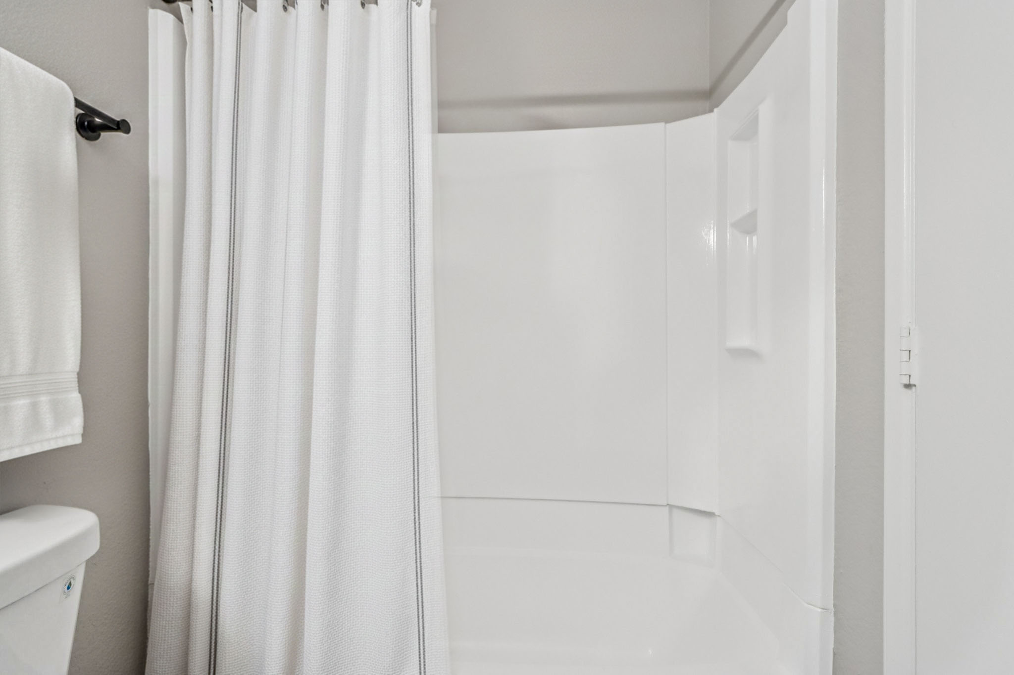 Shower area with a white curtain partially drawn to the side. The walls are light gray, and there is a white toilet visible in the left foreground. The space appears clean and minimalistic, with simple fixtures. A towel hangs on the wall near the shower.