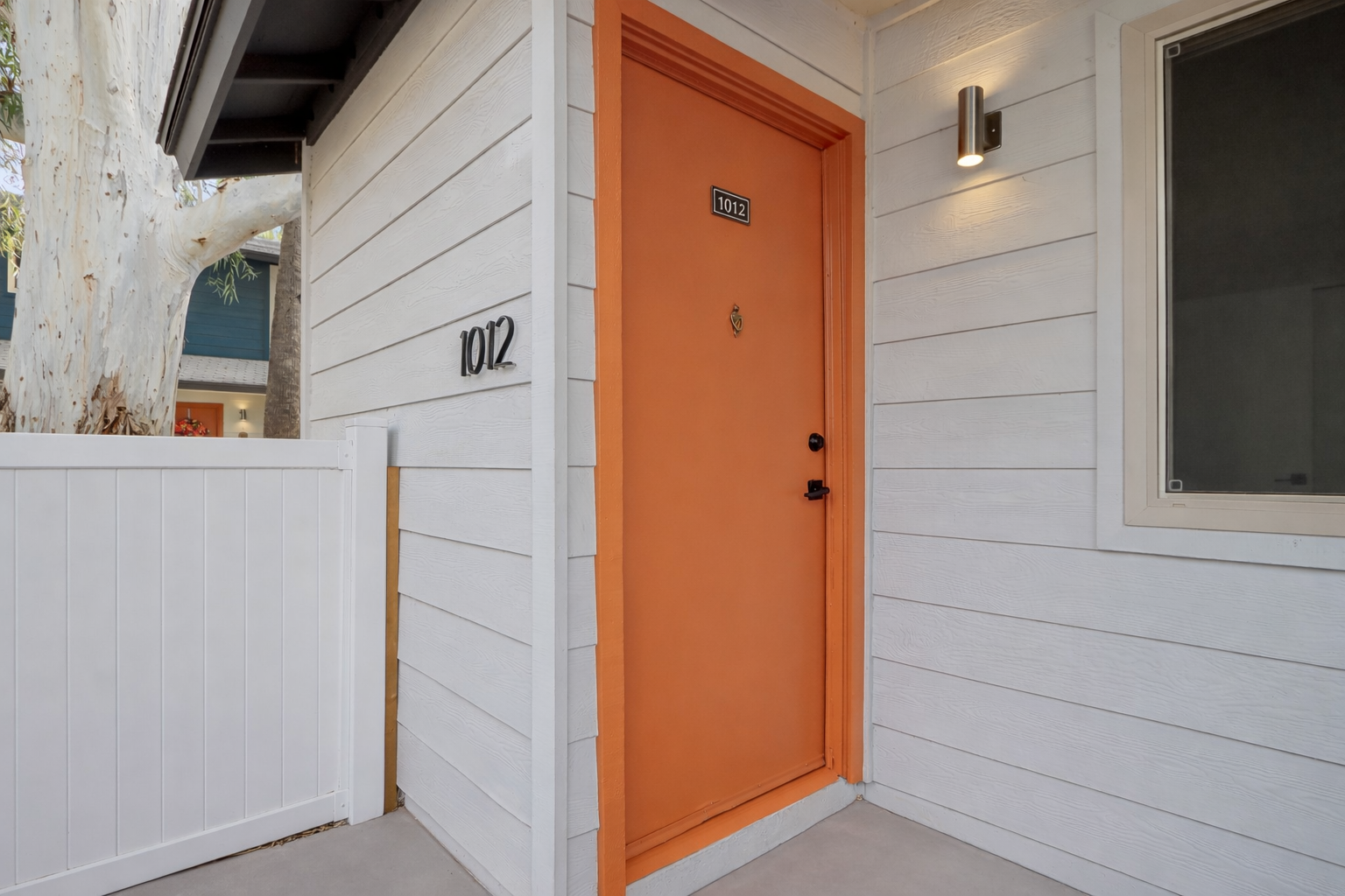 A vibrant orange door marked "1012" on a modern white house exterior, with a light fixture above and a white wooden fence to the left. The entrance leads to a welcoming entryway, framed by neutral-colored siding and large windows that hint at a stylish interior.
