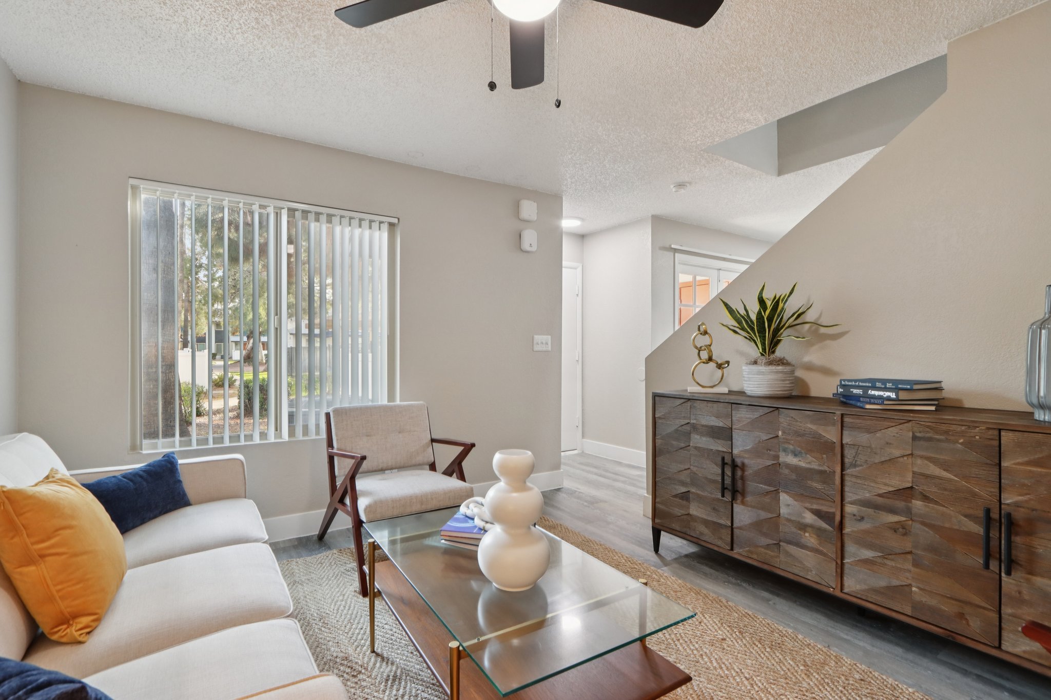 Modern living room featuring a light-colored sofa with blue and orange cushions, a glass coffee table, and a wooden sideboard. The space has neutral walls, wooden flooring, and a ceiling fan. Large windows provide natural light, and decorative elements like plants and books enhance the cozy atmosphere.