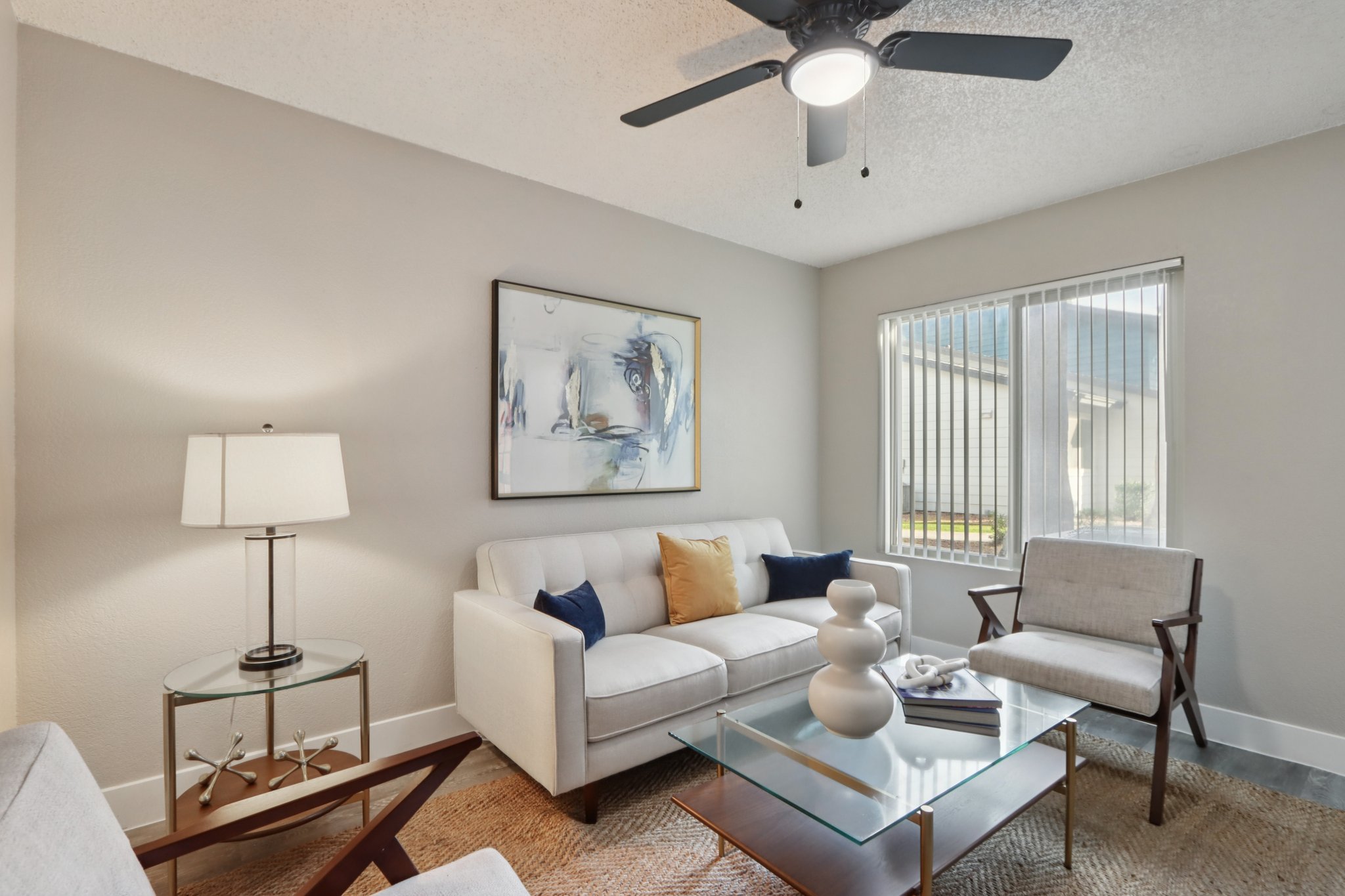 A modern living room featuring a light-colored sofa with accent pillows, a glass coffee table adorned with a decorative vase and a book, and two stylish chairs. A lamp stands beside the sofa, and a painting hangs on the wall. Natural light streams in through a window with blinds.
