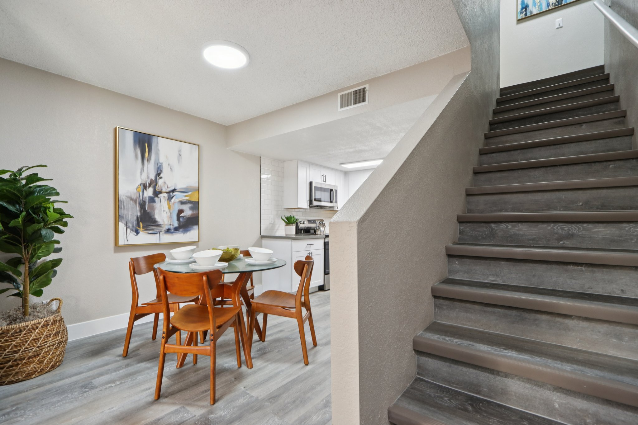 A modern dining area featuring a round glass table set with white plates. Surrounding the table are four wooden chairs. To the side, there is a decorative potted plant. The room has light gray walls and flooring, with a staircase leading to an upper level and an abstract painting on the wall.