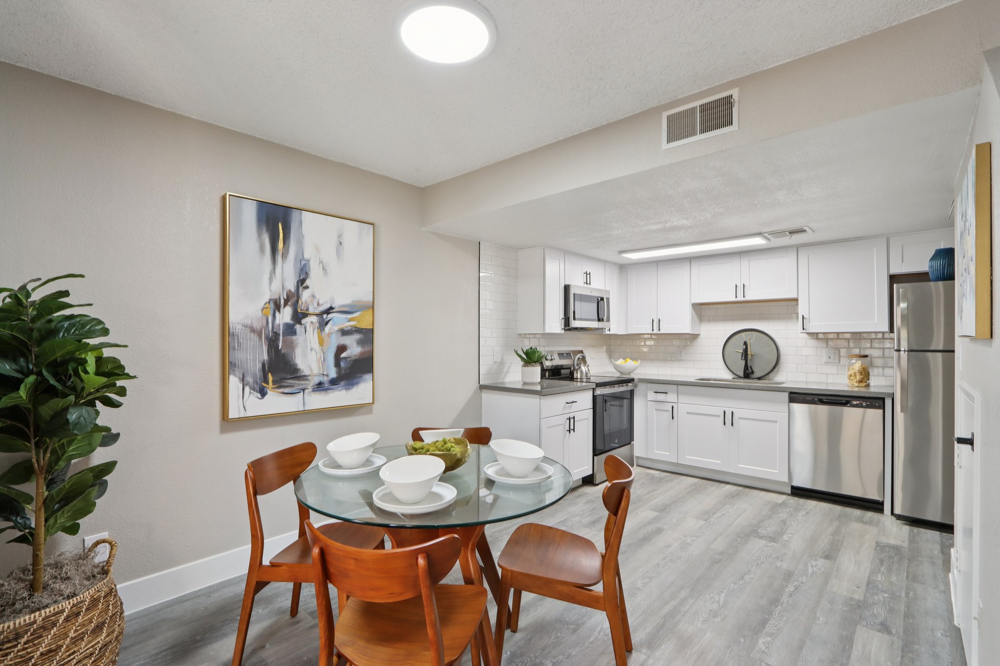 A modern kitchen and dining area featuring a round glass table set with white dishes and wooden chairs. The kitchen has white cabinets, stainless steel appliances, and sleek countertops. A potted plant adds a touch of greenery, while a colorful abstract painting decorates the wall, enhancing the contemporary aesthetic.