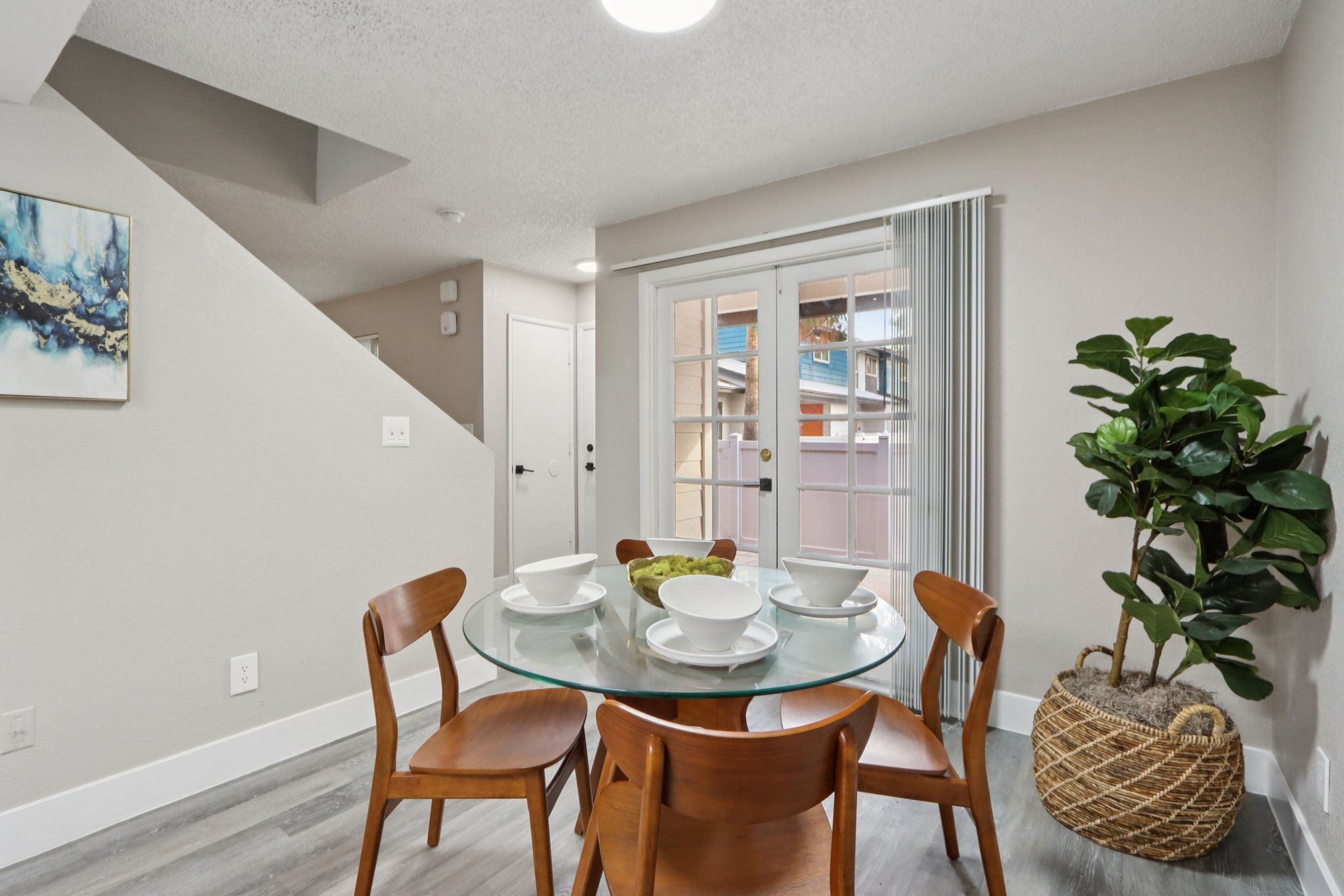 A modern dining area featuring a round glass table set for four with white bowls and a centerpiece. Surrounding the table are wooden chairs. In the background, there are sliding glass doors leading to an outdoor space, and a potted plant adds greenery to the room. The walls are painted a neutral color, enhancing the bright atmosphere.