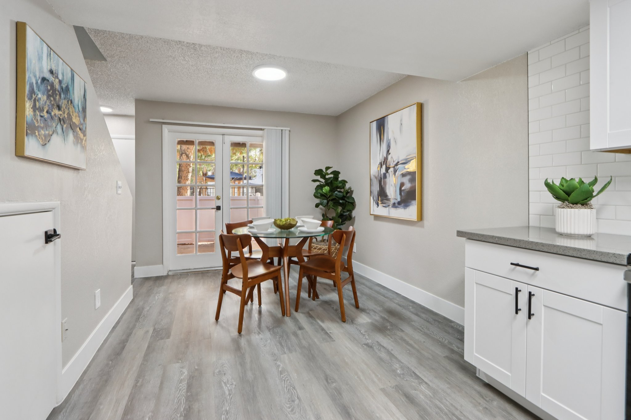 A modern dining area featuring a round table with four wooden chairs, a decorative plant, and artwork on the walls. Natural light enters through sliding glass doors leading to a patio. The space has light-colored walls and sleek gray flooring, creating a warm and inviting atmosphere.