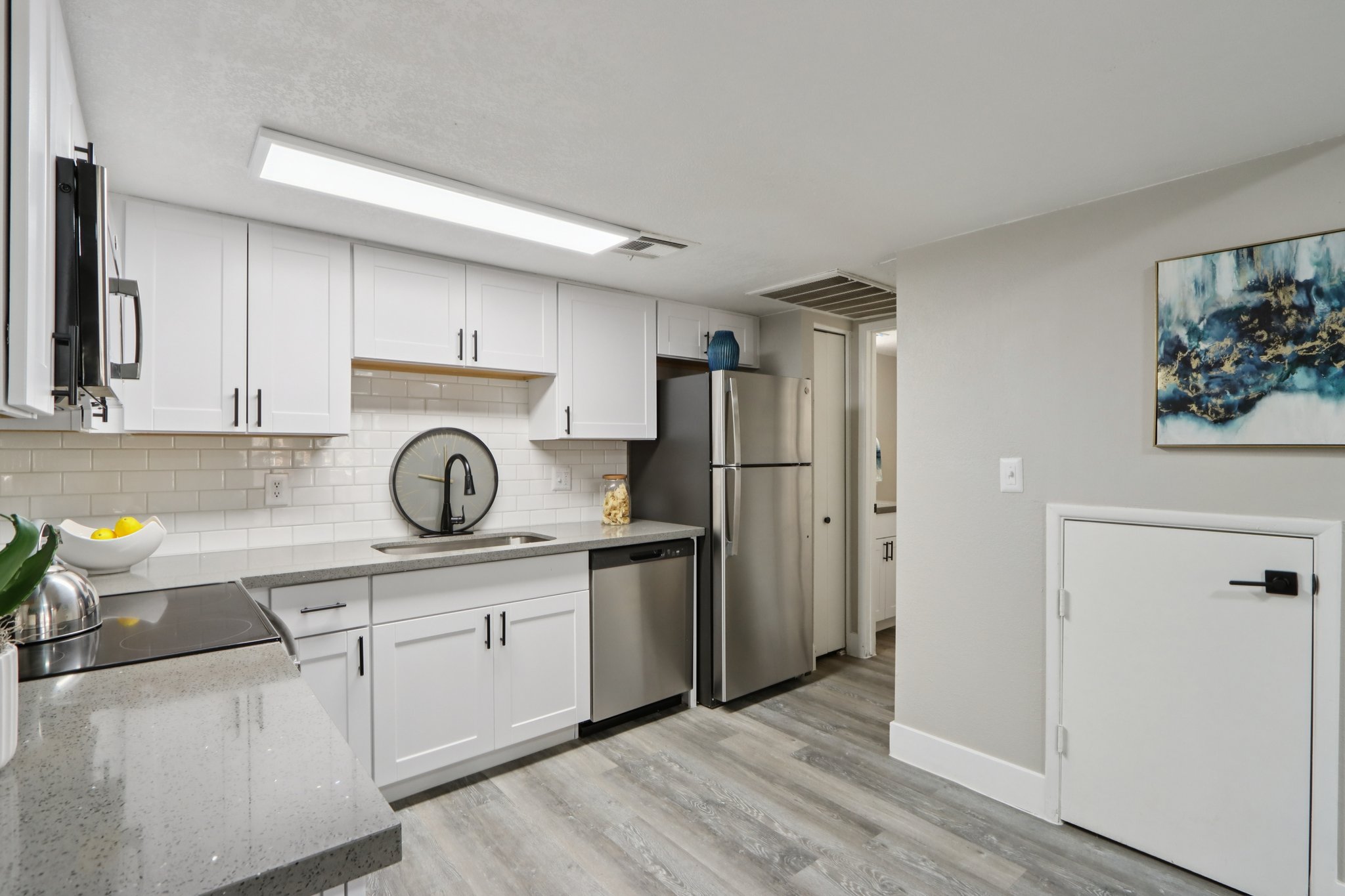 Modern kitchen featuring white cabinets, a stainless steel refrigerator, and a sleek countertop. The design includes a circular mirror above the sink and a decorative bowl of fruit. Light-colored flooring complements the bright and airy atmosphere of the space.