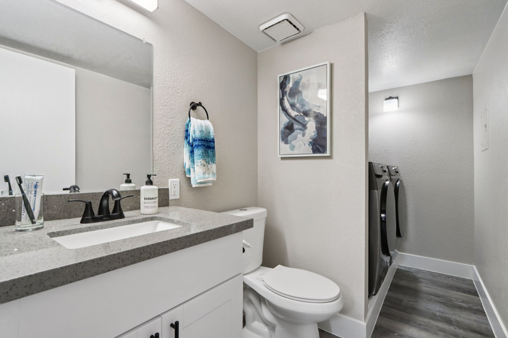 A modern bathroom featuring a gray countertop sink, a white toilet, and a large mirror. The walls are painted light gray, and there's a colorful abstract artwork on the wall. A hand towel hangs nearby, and a laundry area with two black washing machines is visible in the background. The flooring is a dark wood-like material.