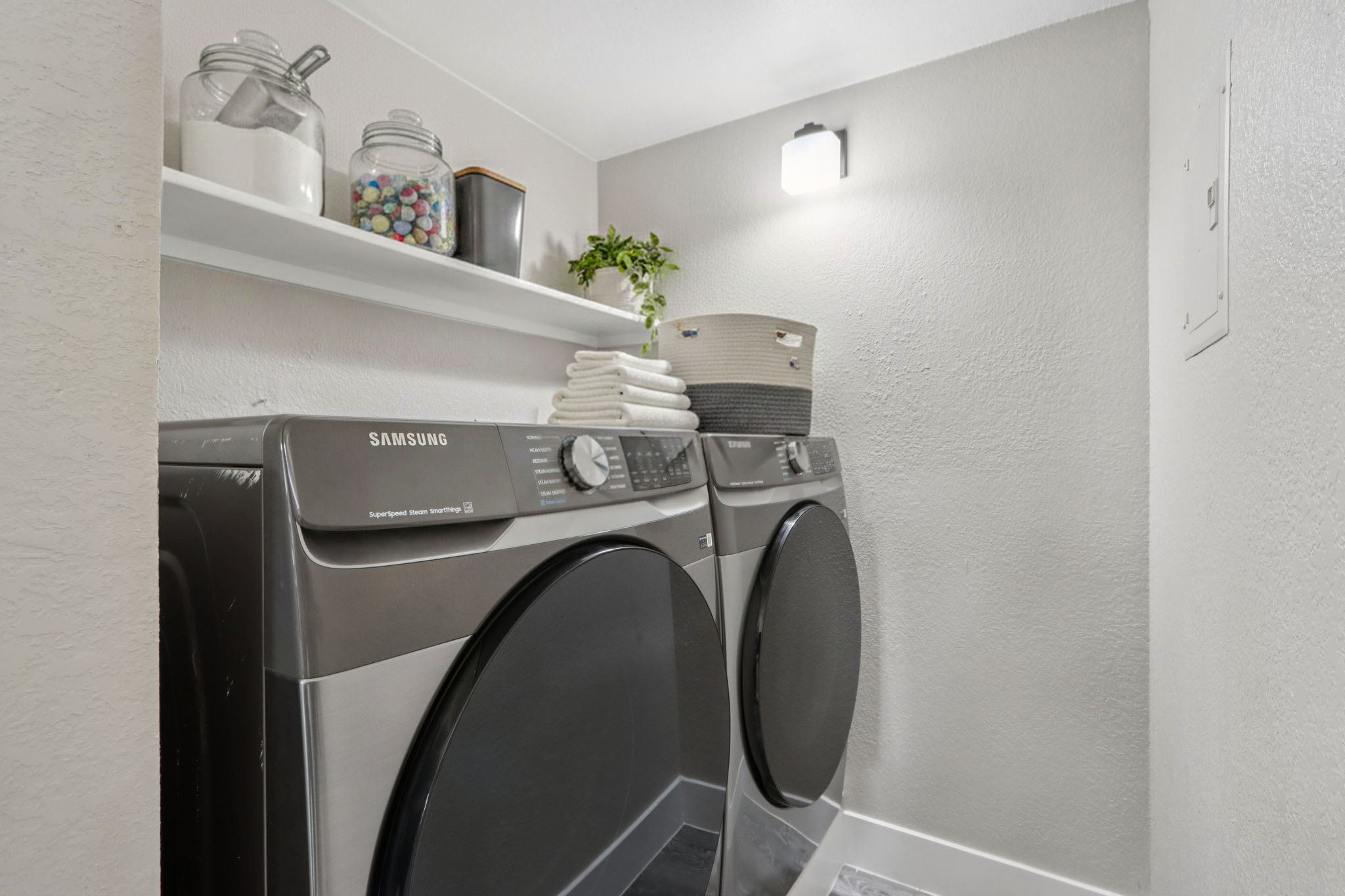 A modern laundry room featuring a pair of Samsung washing machines and a drying machine. Above, a shelf displays jars and decorative items, while neatly stacked towels and a woven basket sit nearby. The walls are painted in a light color, and a wall-mounted light fixture provides illumination.