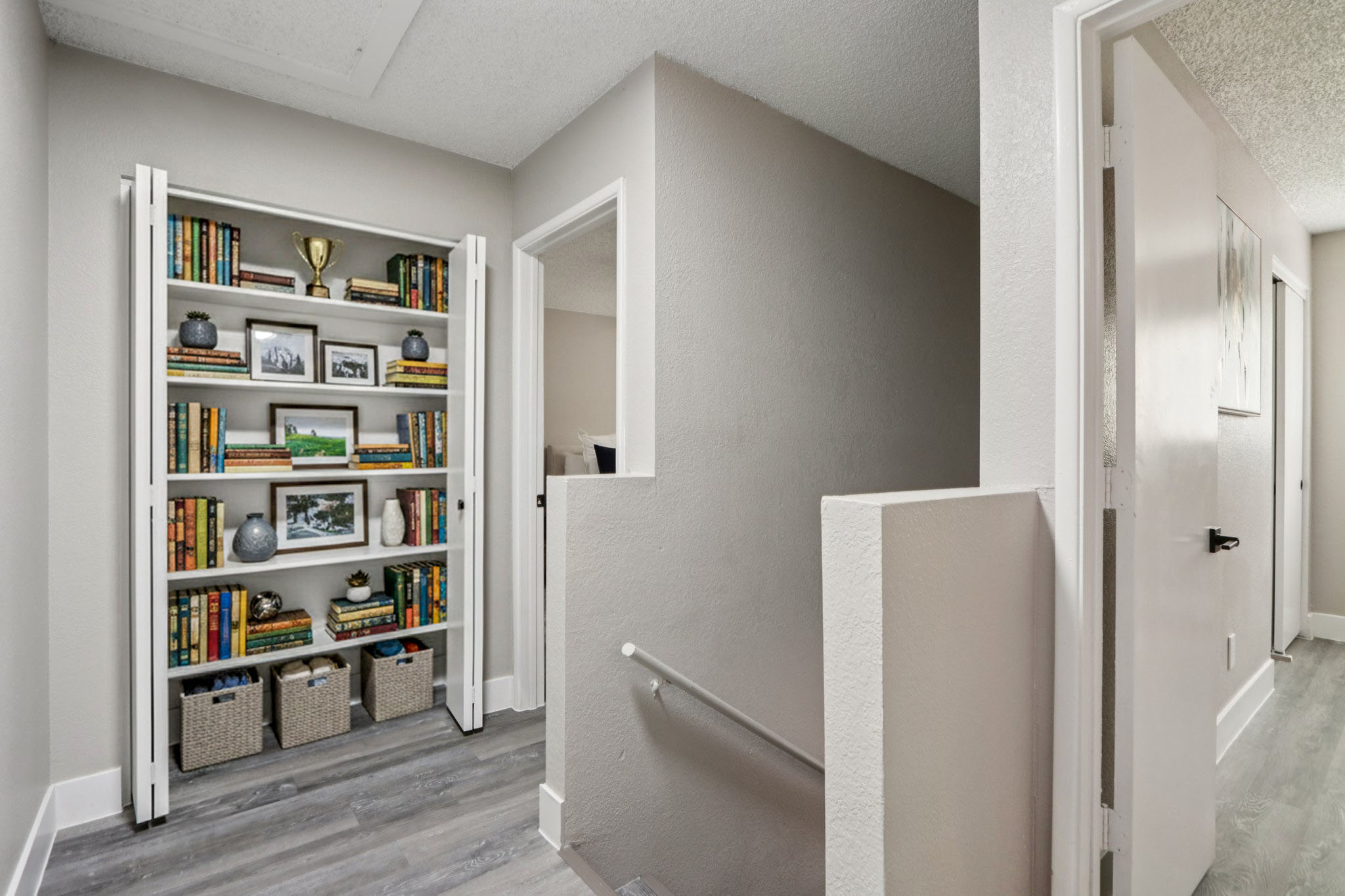 A well-lit hallway featuring a built-in bookshelf filled with colorful books and decorative items. There are two doorways on the right, and the floor is a light wood laminate. The walls are painted a soft gray, creating a modern and inviting atmosphere.