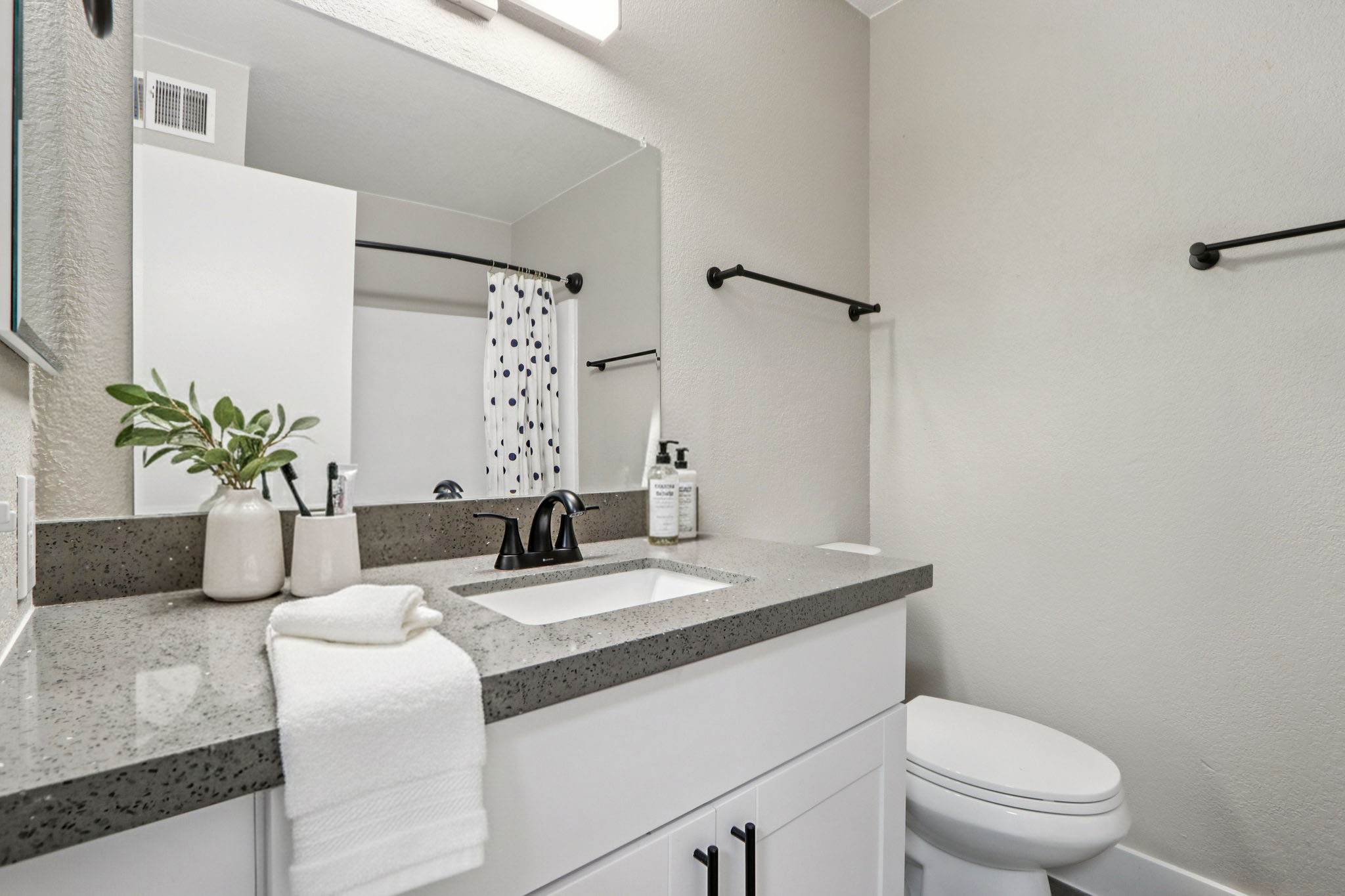 A clean and modern bathroom featuring a gray countertop with a white sink, a stylish faucet, and neatly arranged toiletries. There is a white towel rolled on the counter, a small plant in a white vase, and a large mirror above the sink. The wall is painted in a light color, with a shower curtain visible in the background.