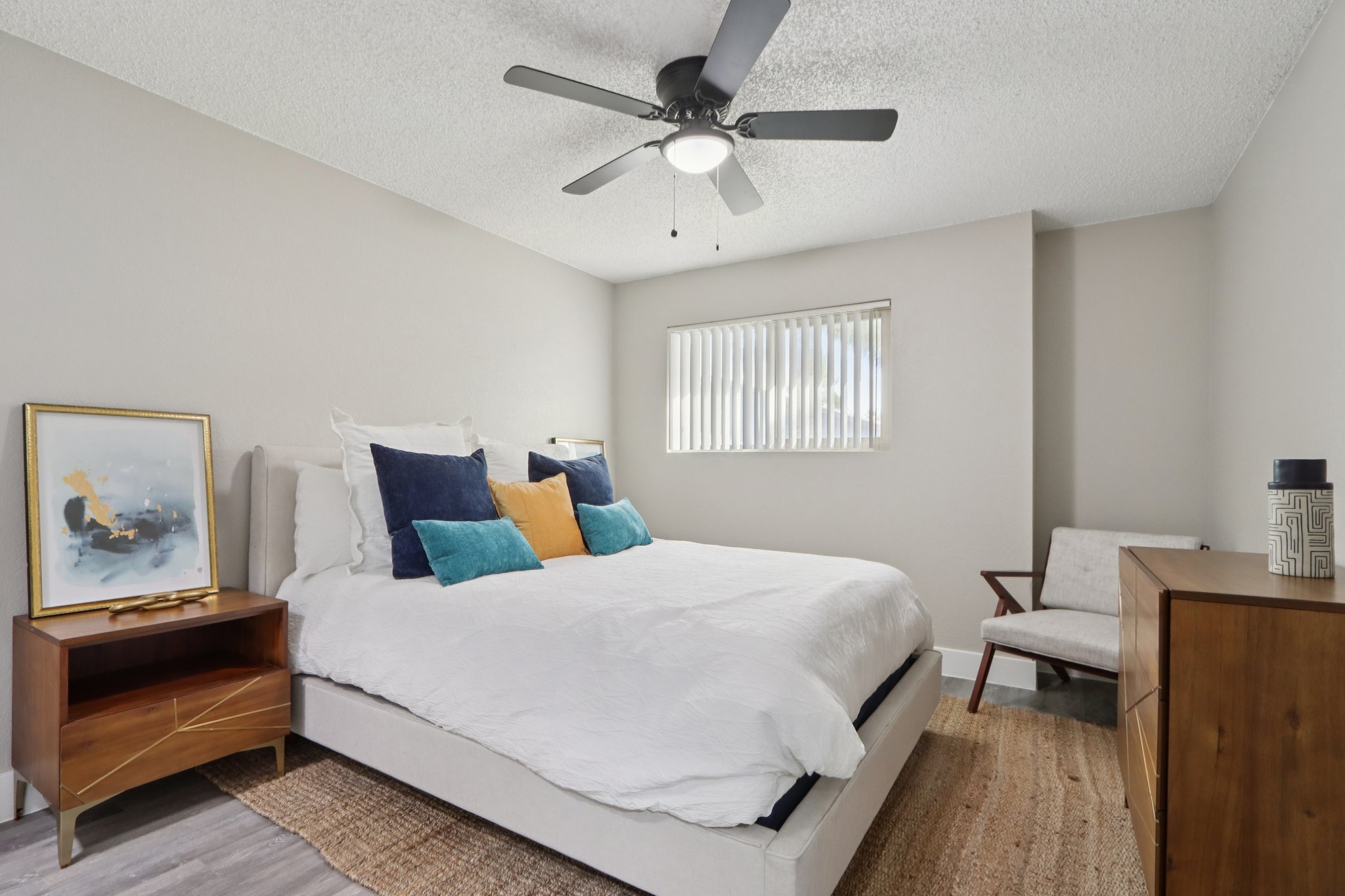 A cozy bedroom featuring a queen-sized bed with white bedding and accent pillows in blue and orange. There's a modern ceiling fan and a large window with vertical blinds, allowing natural light to enter. A wooden nightstand and a chair complement the decor, along with an artistic framed picture on the wall.
