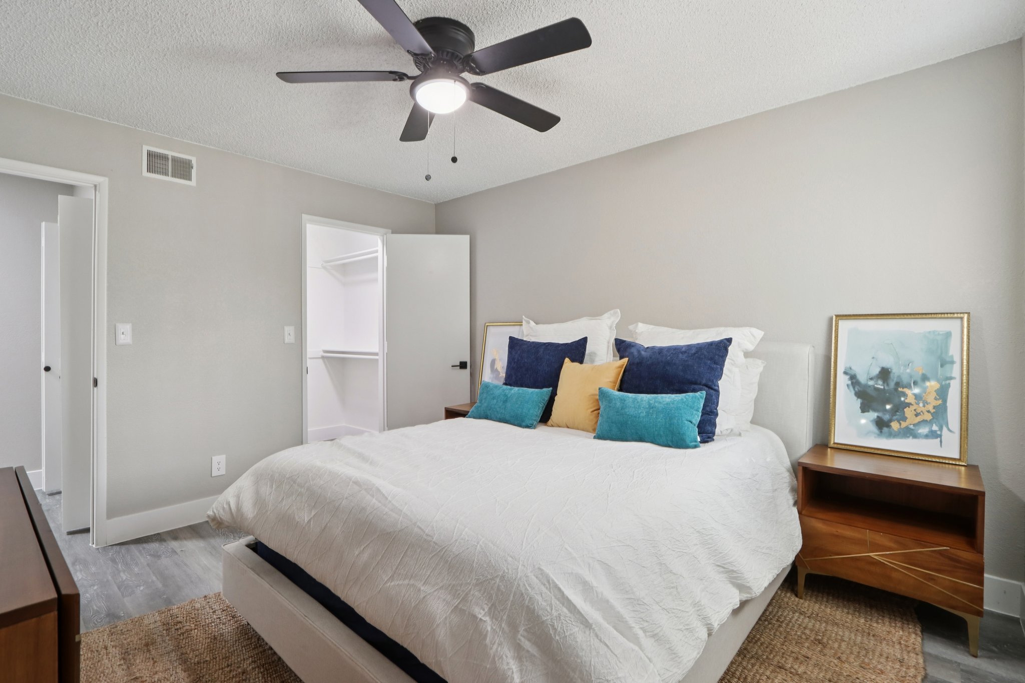 A cozy bedroom featuring a white bed with decorative pillows in blue and yellow, a ceiling fan, and wooden nightstands. A closet door is partially open in the background, and there are light gray walls and a textured rug on the floor, giving a clean and inviting atmosphere.