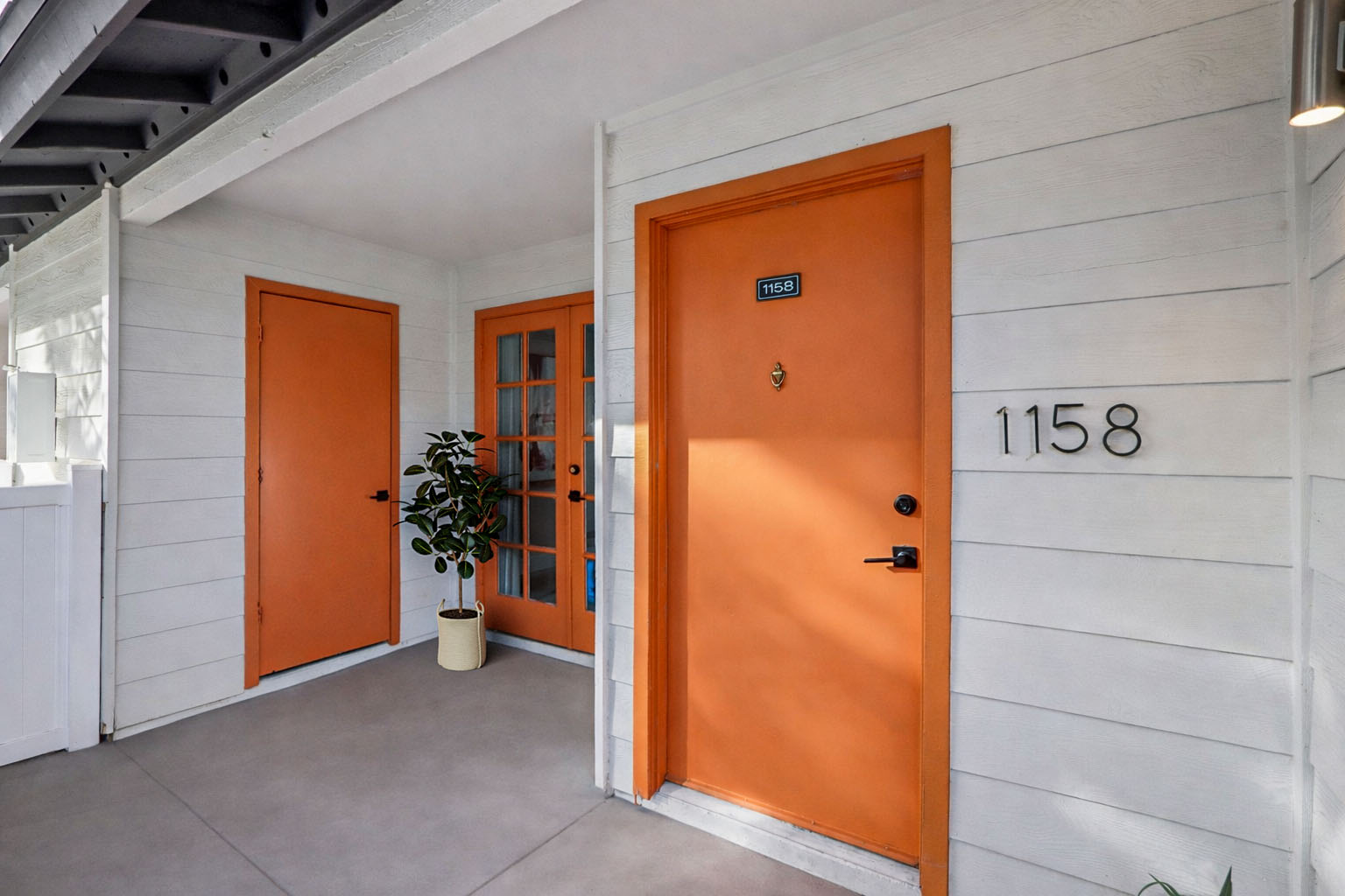 Exterior view of a residential entryway featuring a white wooden facade with two orange doors. The door on the left is a standard door, while the one on the right has glass panels. A small potted plant is positioned near the wall beside the doors. The number "1158" is displayed on the wall next to the door.