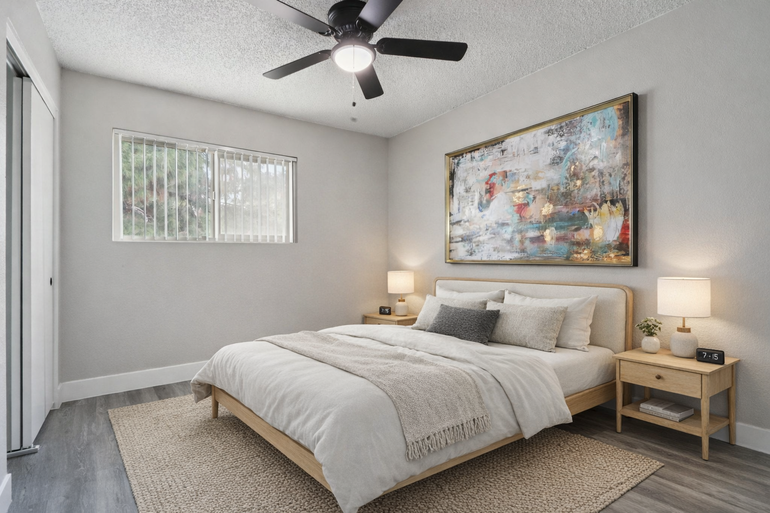 A modern bedroom featuring a queen-sized bed with neutral-toned bedding, two bedside lamps, and a wooden nightstand. A large abstract painting is above the bed, and the room has light gray walls and a ceiling fan. Natural light filters through a window with blinds. A textured area rug covers the floor.