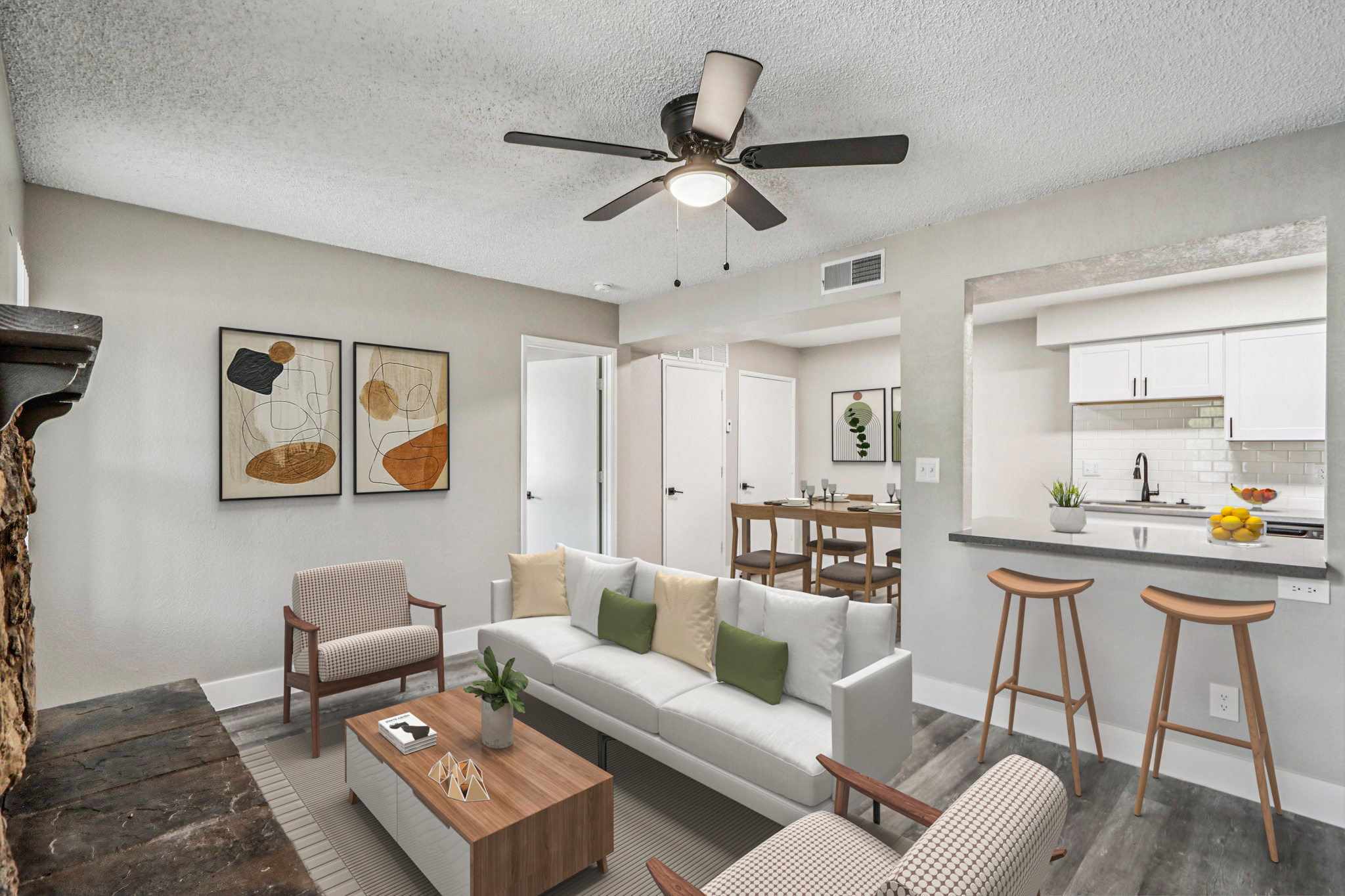 A modern living room with a white sofa adorned with green and beige pillows, two light armchairs, a wooden coffee table, and decorative items. A ceiling fan is visible, along with framed abstract art on the walls. In the background, a dining area and kitchen can be seen, featuring a clean, contemporary design.