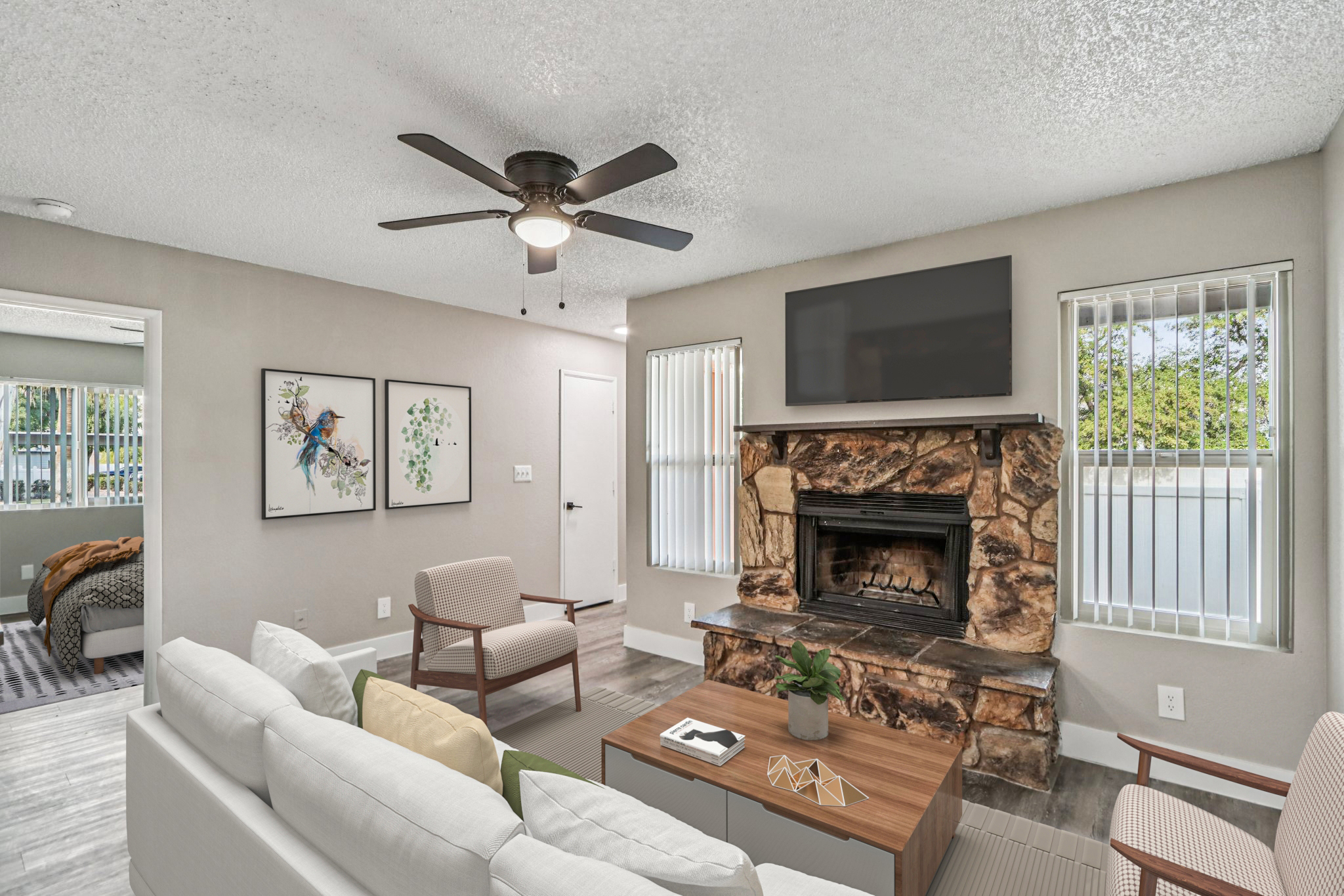 A cozy living room featuring a white sectional sofa, a wooden coffee table, and a stone fireplace with a mounted TV above it. The room has a ceiling fan, natural light from the windows, and two framed artworks on the wall. A doorway leads to another room in the background with a patterned bedspread visible.