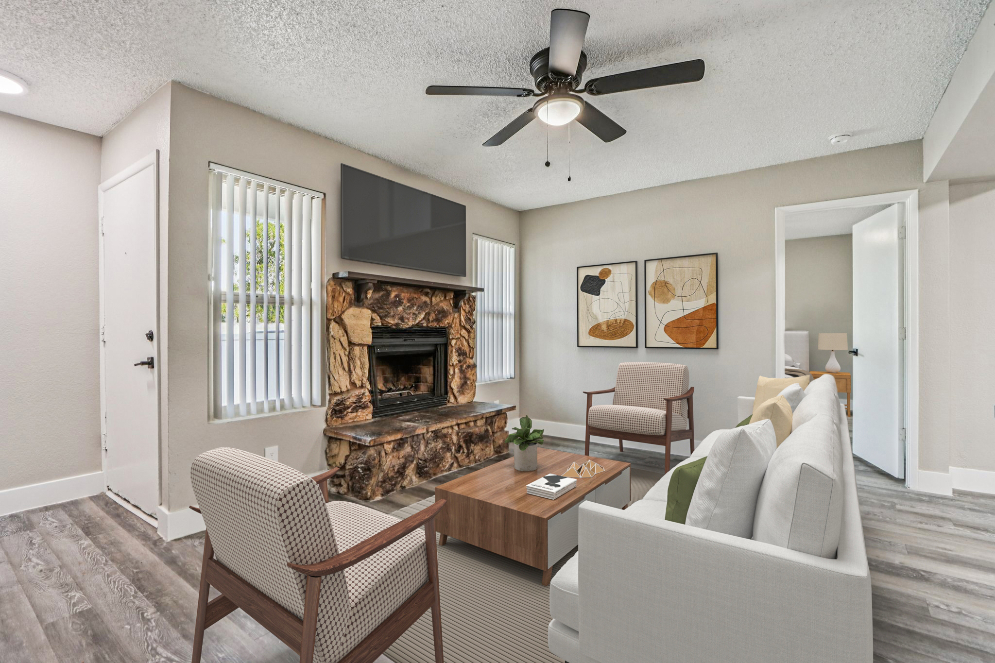 A modern living room featuring a white sofa and two armchairs, a wooden coffee table, and a stone fireplace. There is a ceiling fan above, large windows with blinds, and contemporary artwork on the walls. The space is well-lit and showcases a cozy, stylish atmosphere with neutral colors and sleek furnishings.
