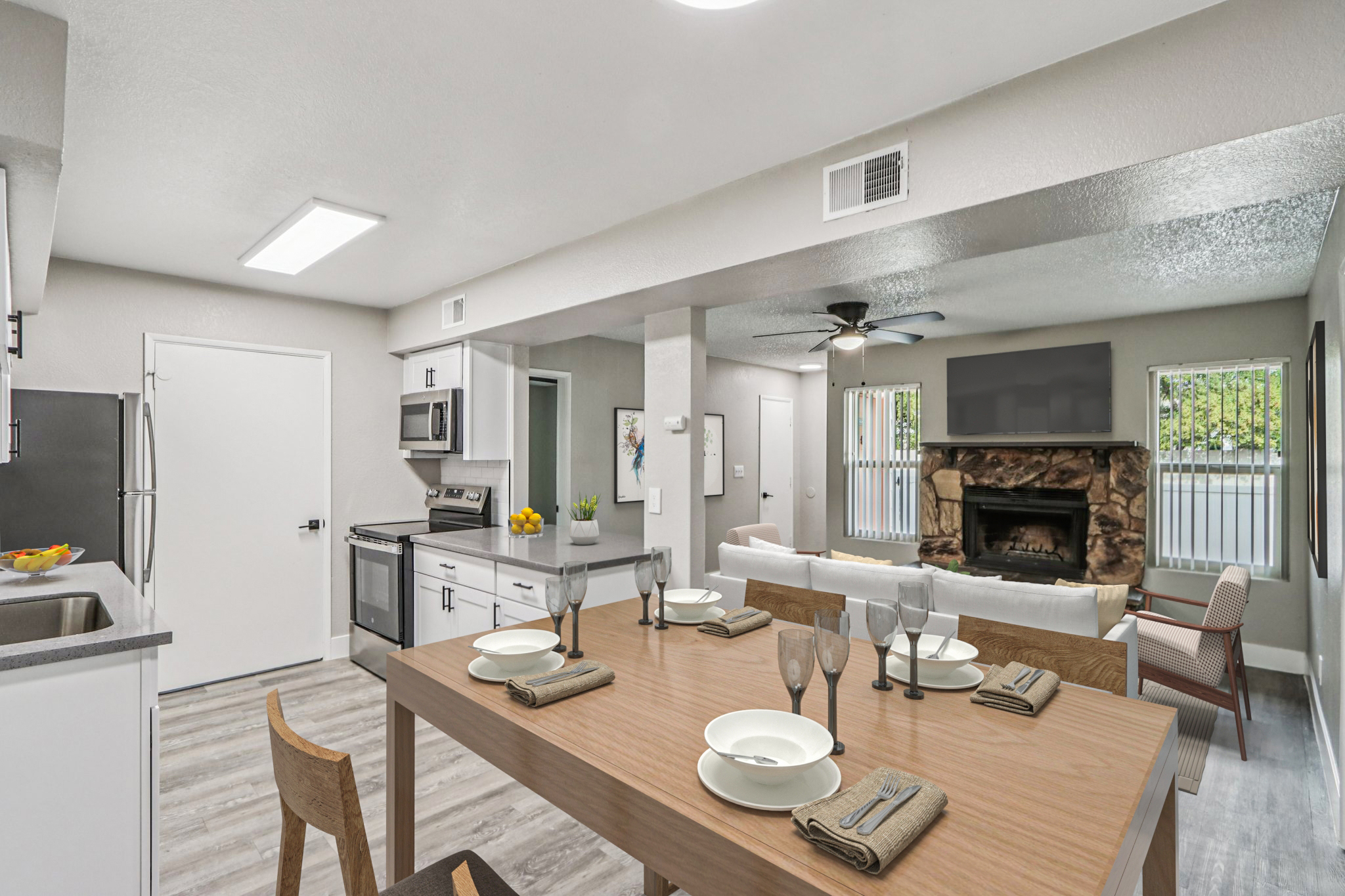 Modern open-concept kitchen and living area featuring a dining table set for six, stainless steel appliances, a cozy stone fireplace, and comfortable seating. Large windows allow natural light to brighten the space, enhancing the warm and inviting atmosphere.
