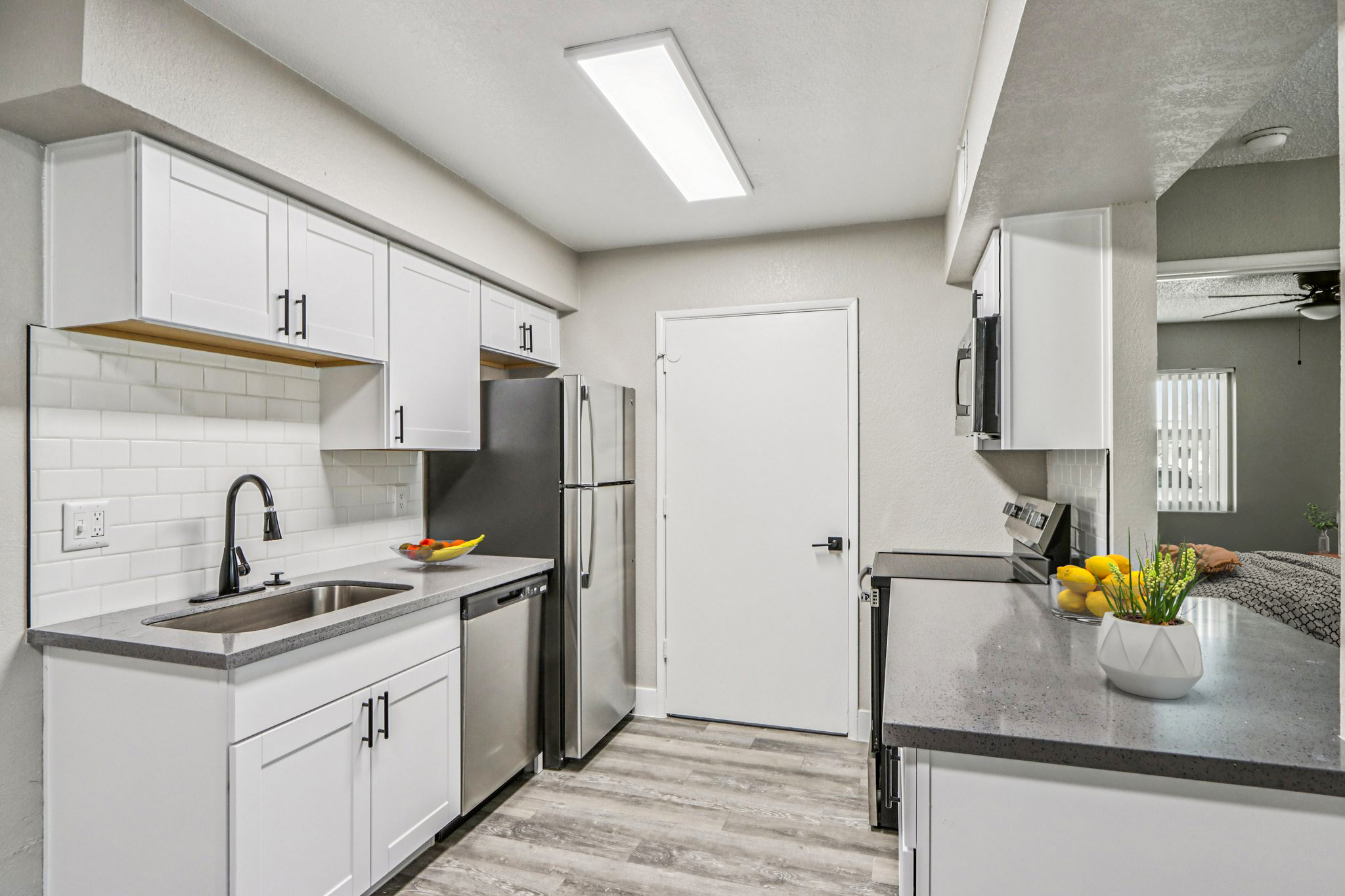 Modern kitchen featuring white cabinets, stainless steel appliances, and a sleek countertop. There is a fruit bowl on the counter, a door leading to another area, and light wood flooring. The overall design is clean and contemporary, with ample natural light.