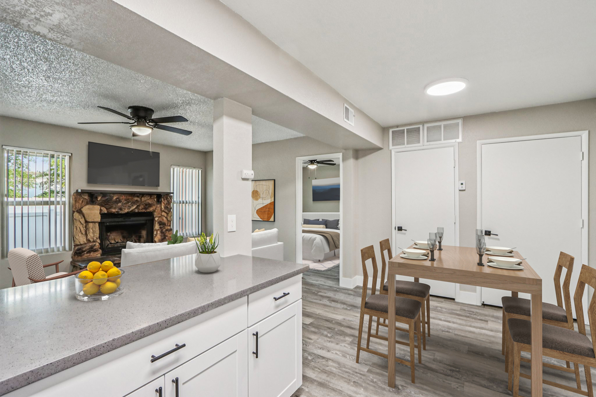 A modern kitchen with gray countertops and a dining area featuring a wooden table set for four. The space is bright and airy, with a stone fireplace in the background, a ceiling fan, and stylish decor, including a bowl of lemons on the counter, large windows, and an inviting atmosphere.