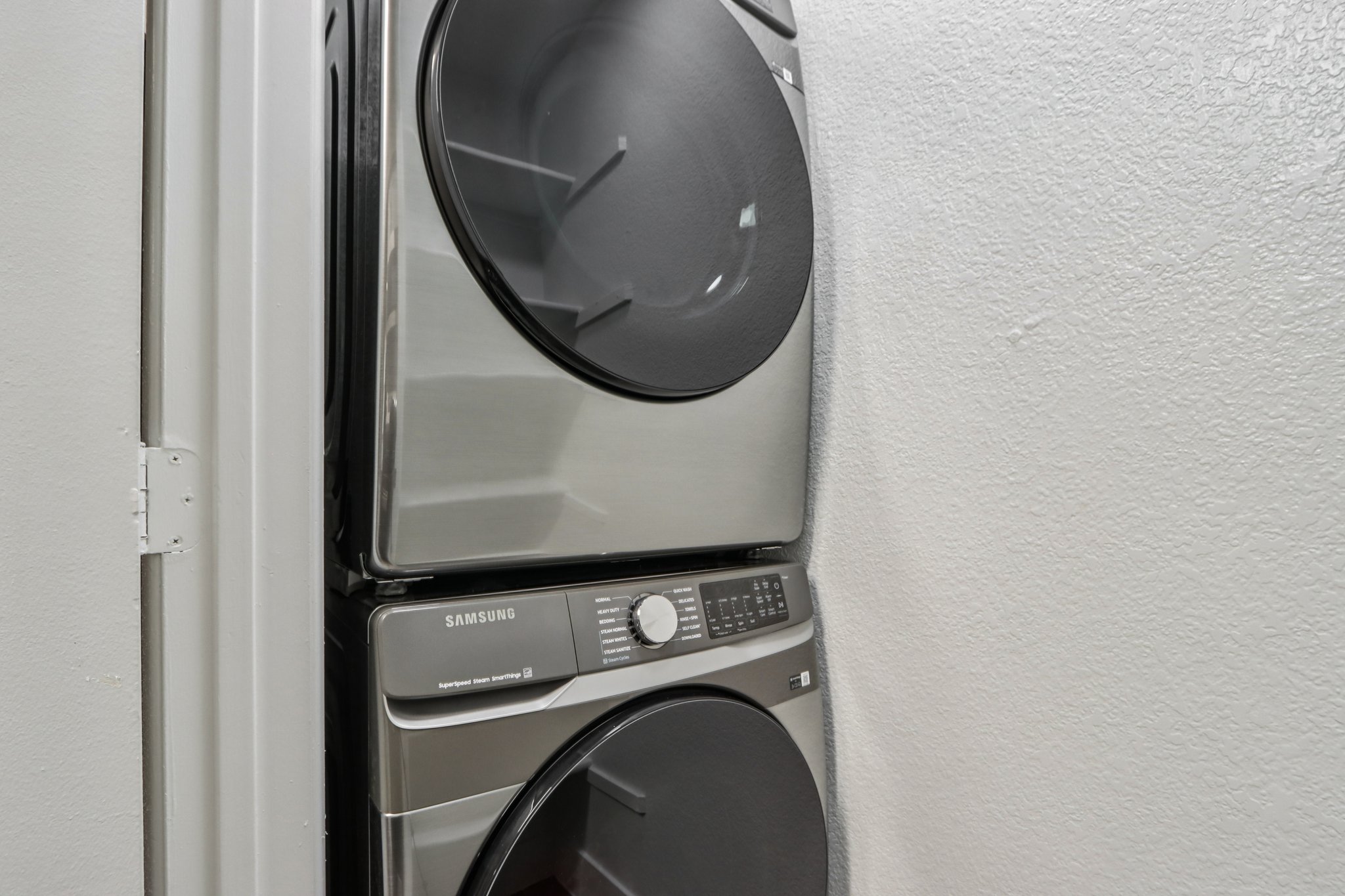 A modern stacked washer and dryer set in stainless steel finish, situated against a white wall. The appliances feature a digital control panel and large circular doors, showcasing a sleek and contemporary design suitable for a laundry room.